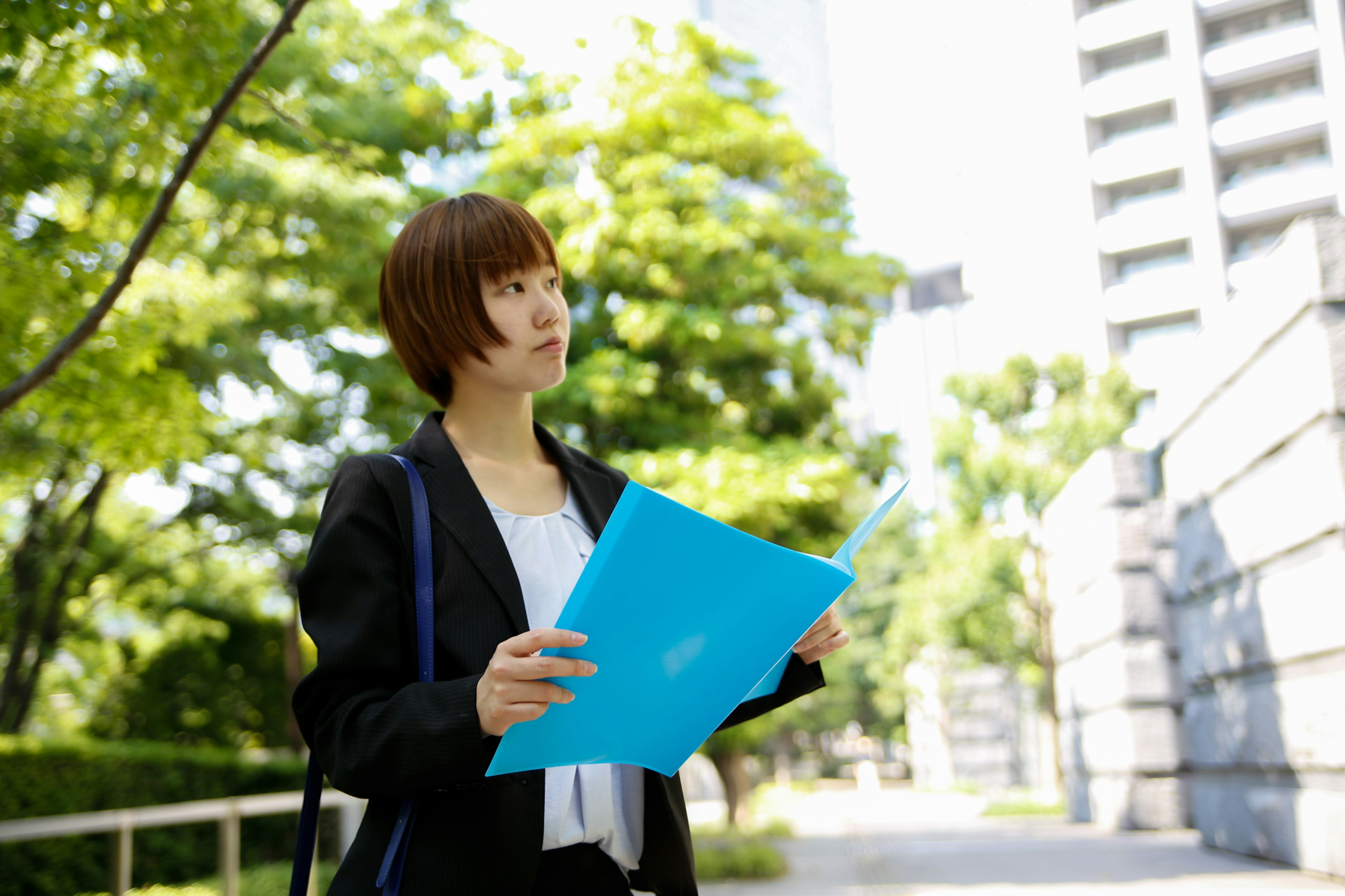 A woman holding a blue folder walking on a path with green trees and tall buildings in the background