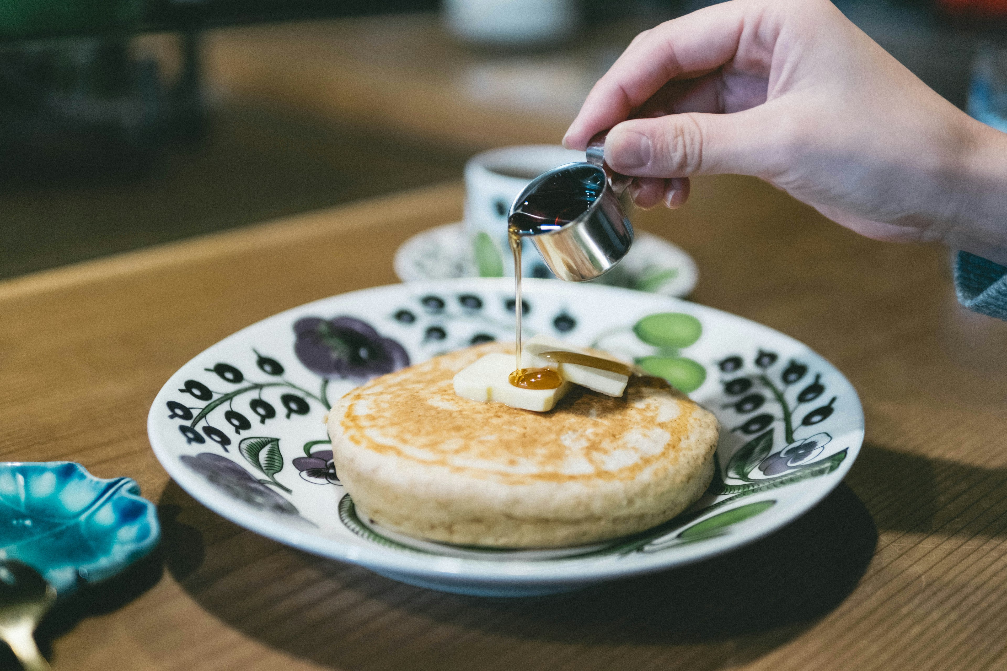 Hand pouring maple syrup over a pancake on a decorative plate