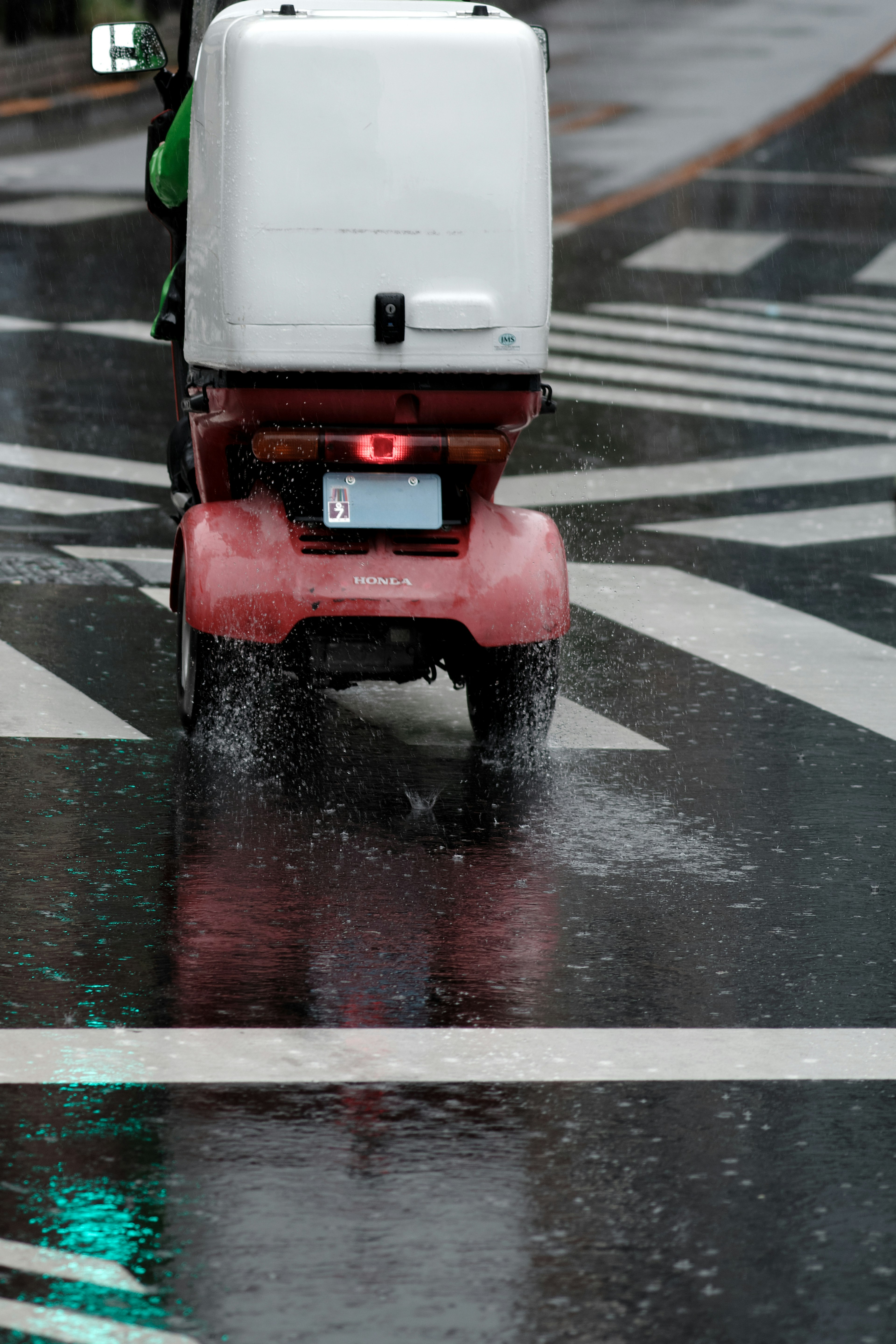 雨に濡れた道路を走る赤い配達スクーターの後ろ姿