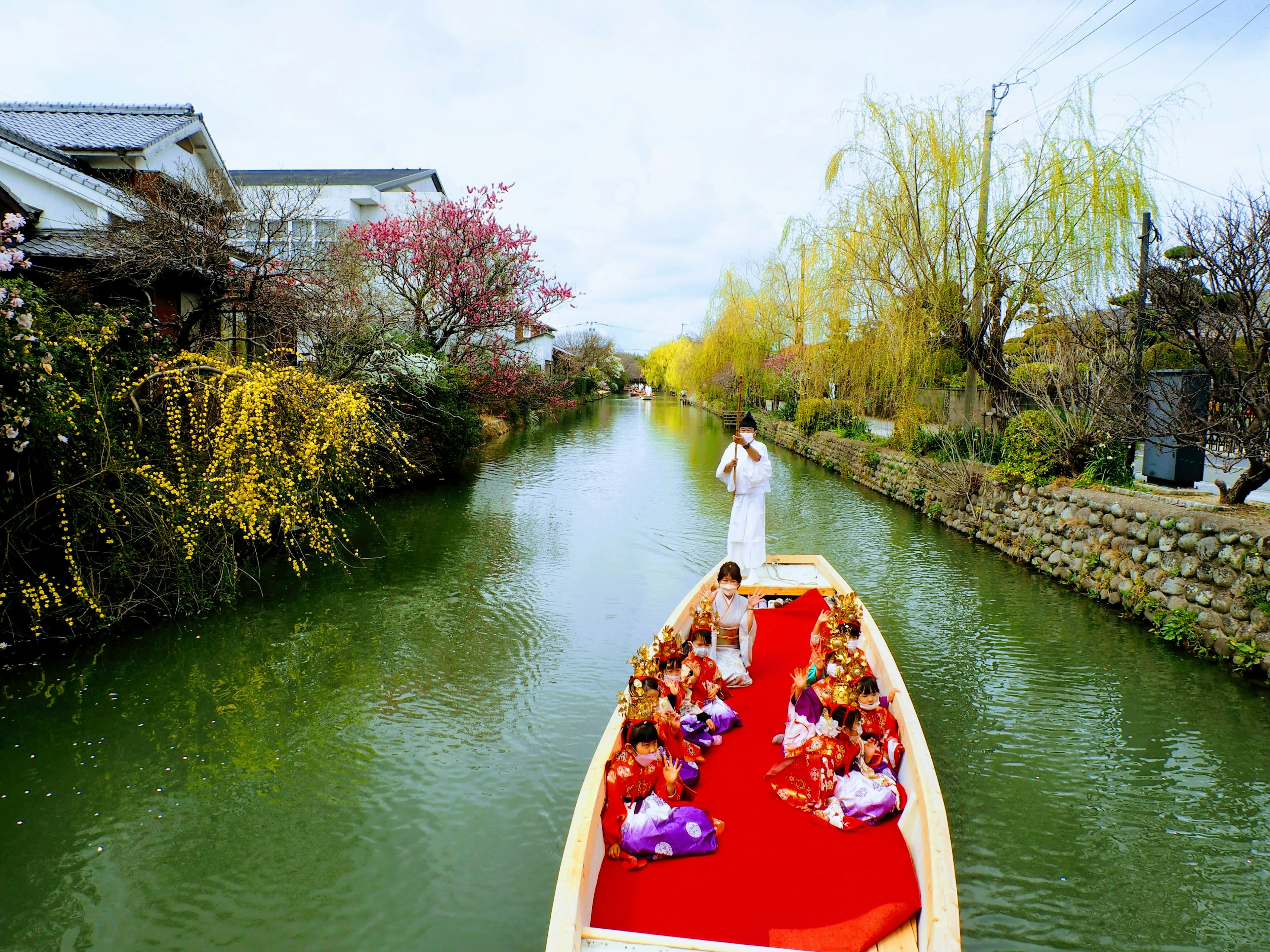 เรือที่มีผู้คนในชุดดั้งเดิมอยู่ในคลองที่เงียบสงบล้อมรอบด้วยดอกไม้ที่บานสะพรั่ง