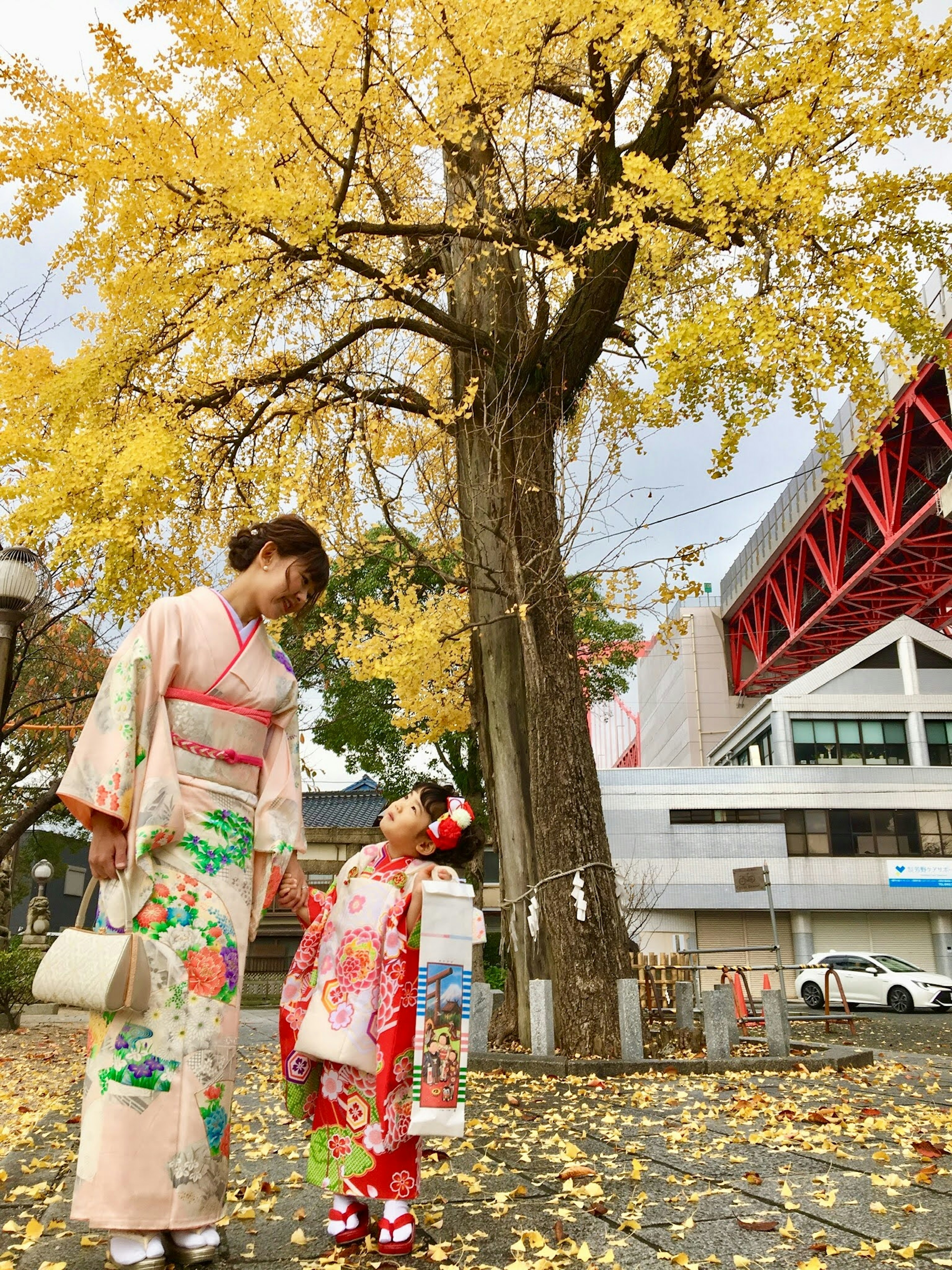 Một người phụ nữ và một đứa trẻ trong trang phục kimono truyền thống đang nhìn nhau dưới một cây ngân hạnh với lá vàng rực rỡ