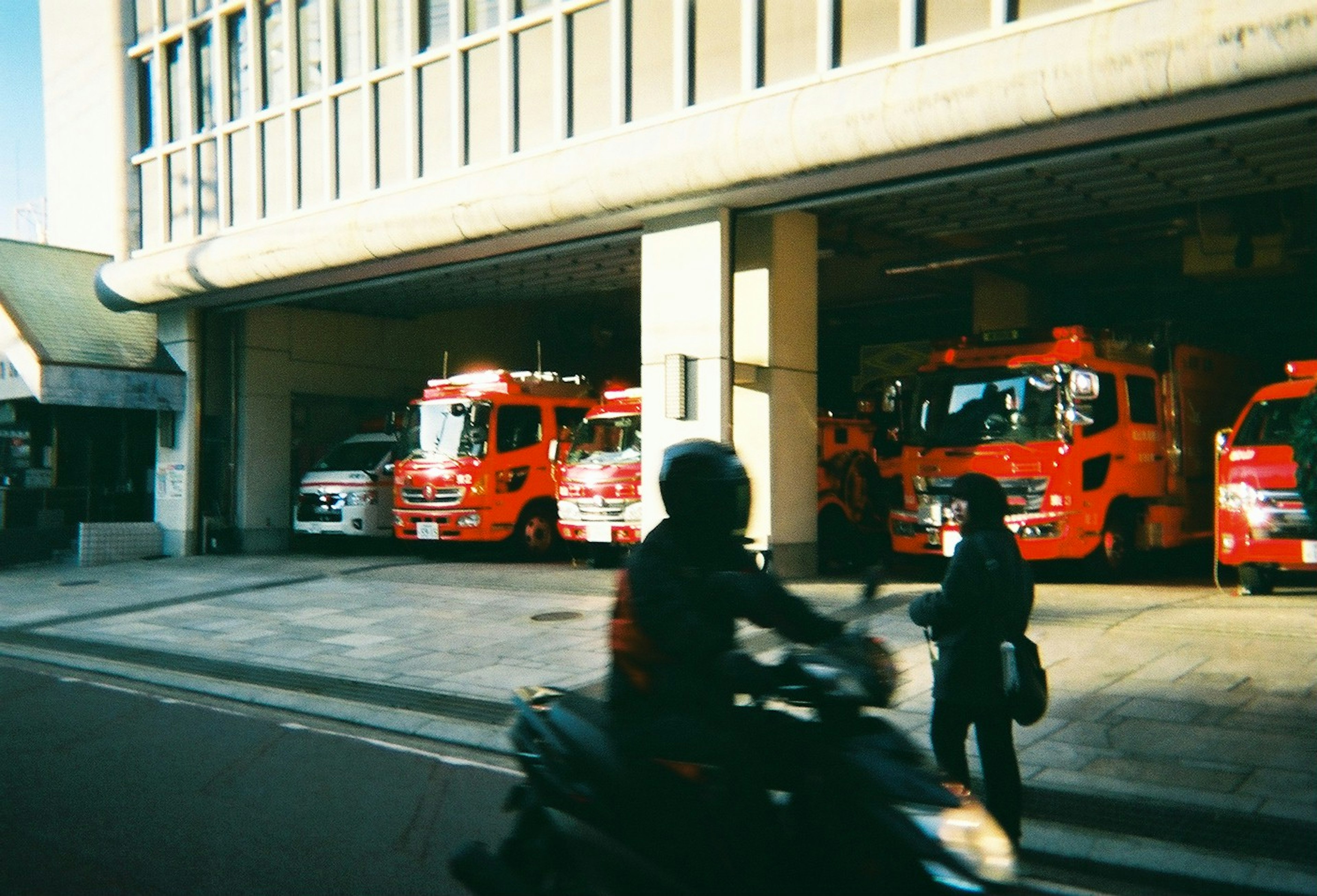 Außenansicht einer Feuerwache mit roten Feuerwehrfahrzeugen und vorbeigehenden Passanten