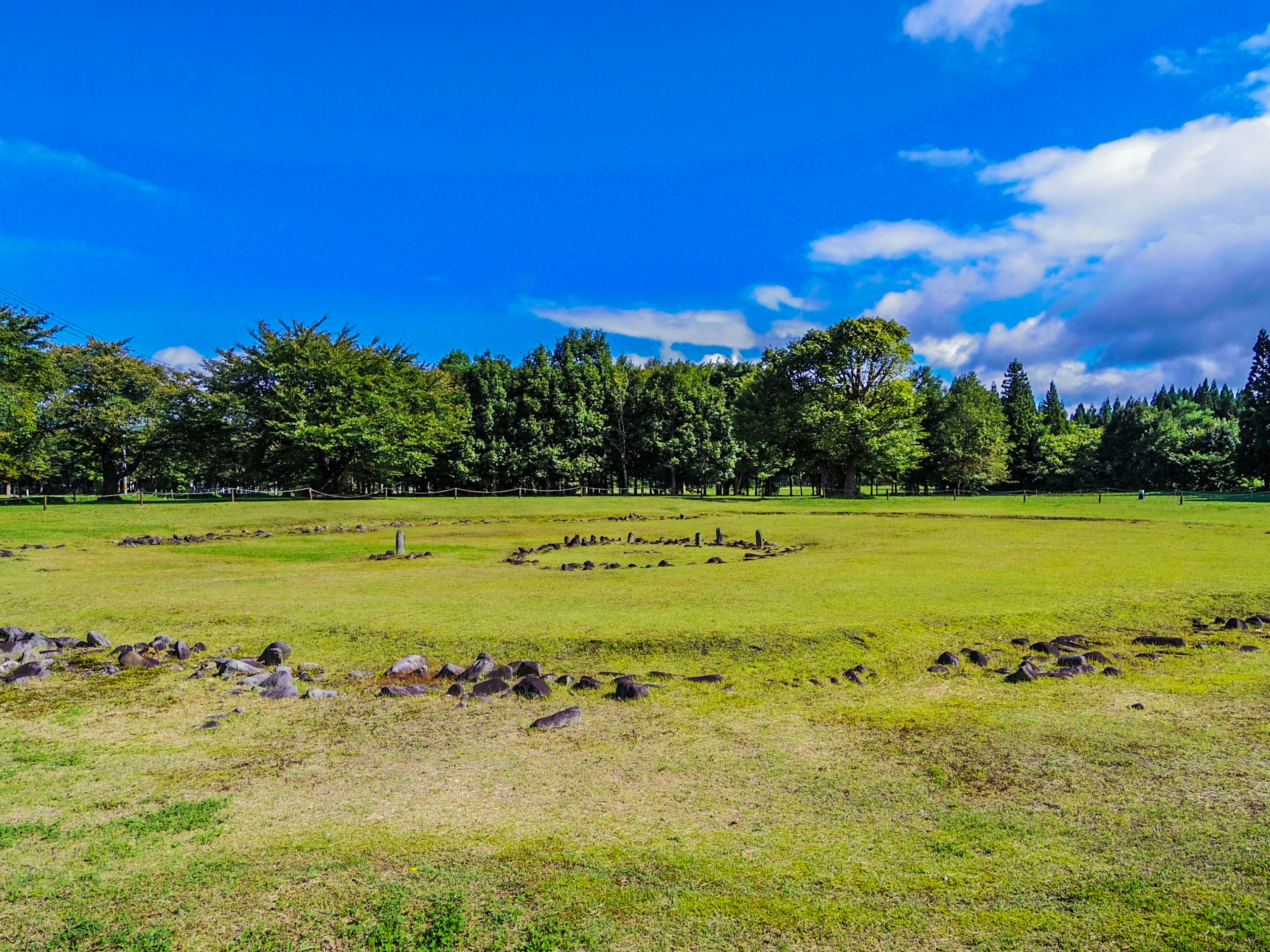 Green meadow under a blue sky featuring circular stone arrangement and lush trees