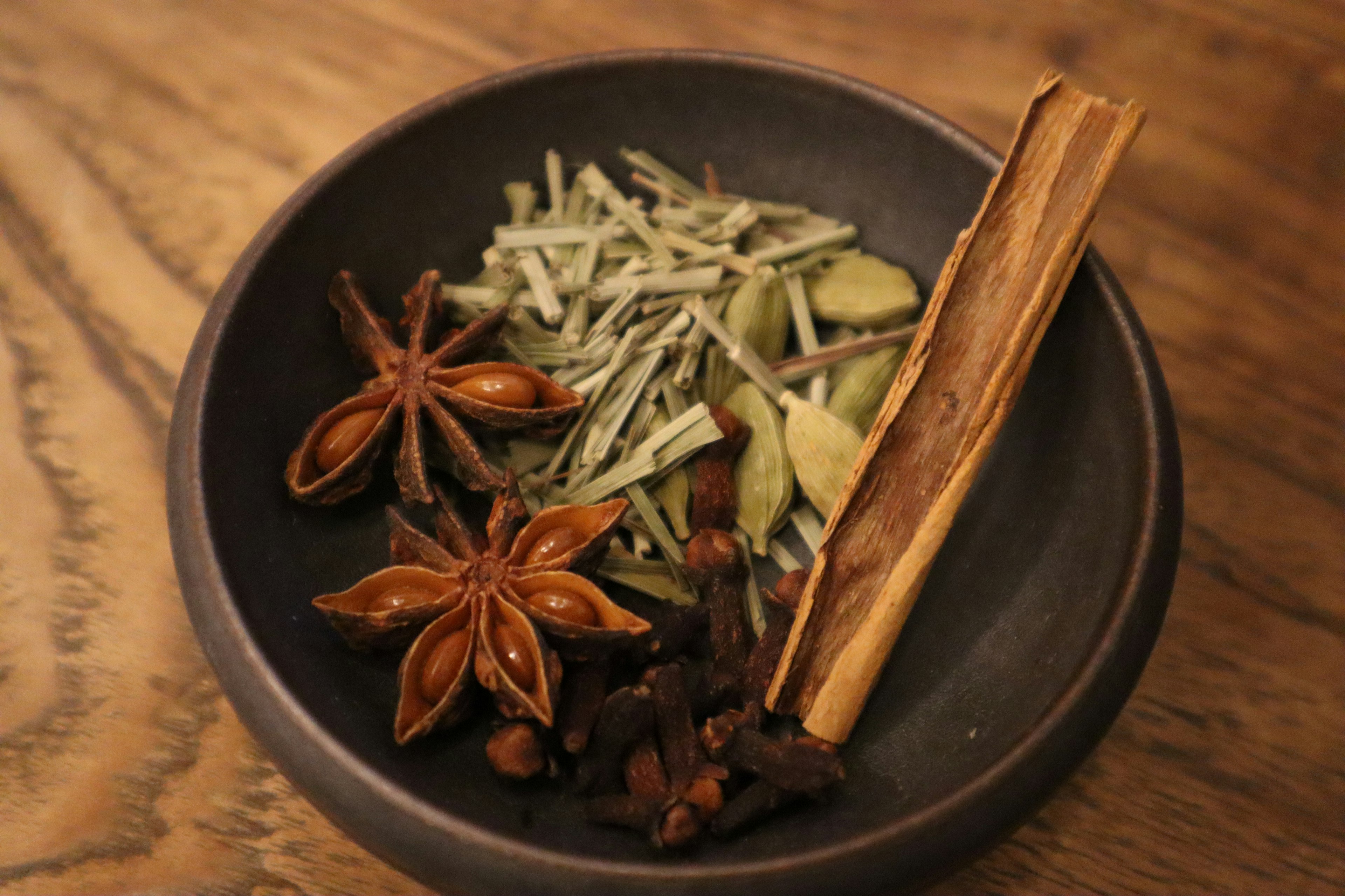 Spices including cinnamon stick, star anise, cloves, lemongrass, and ginger in a black bowl