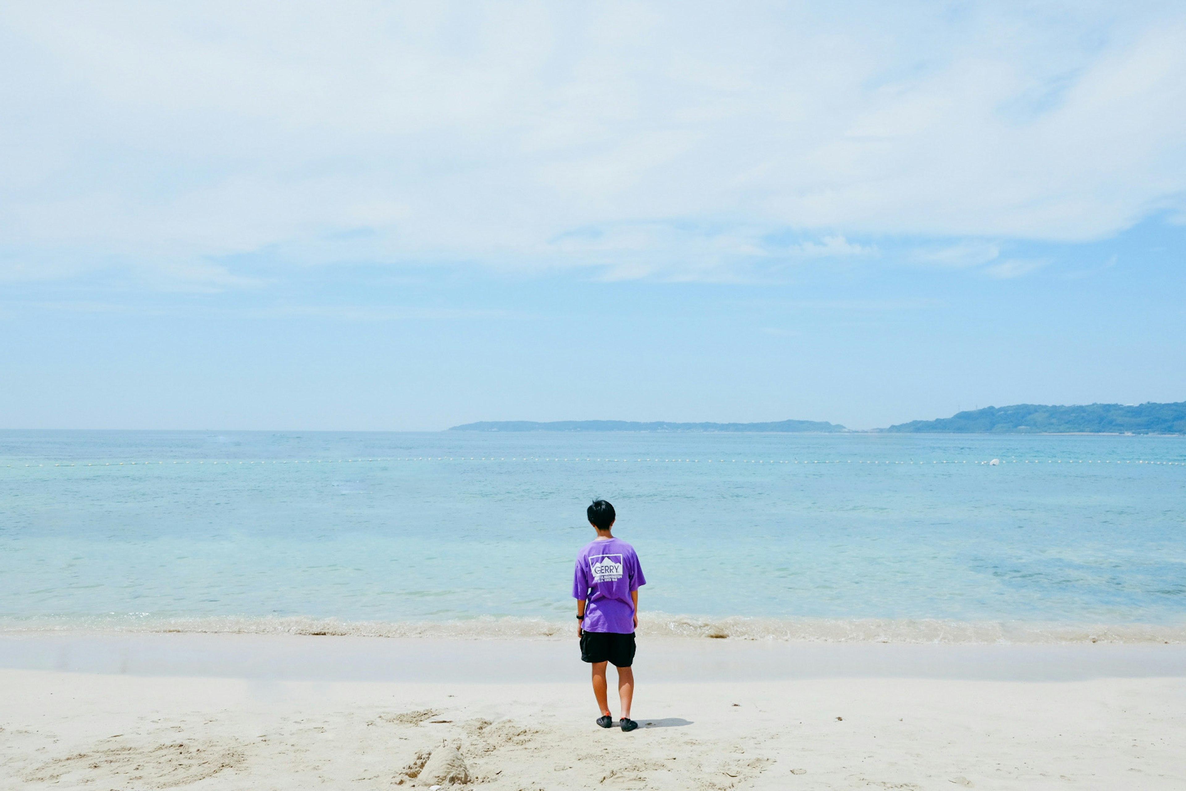 Person in lila T-Shirt steht am Strand und schaut auf den Ozean