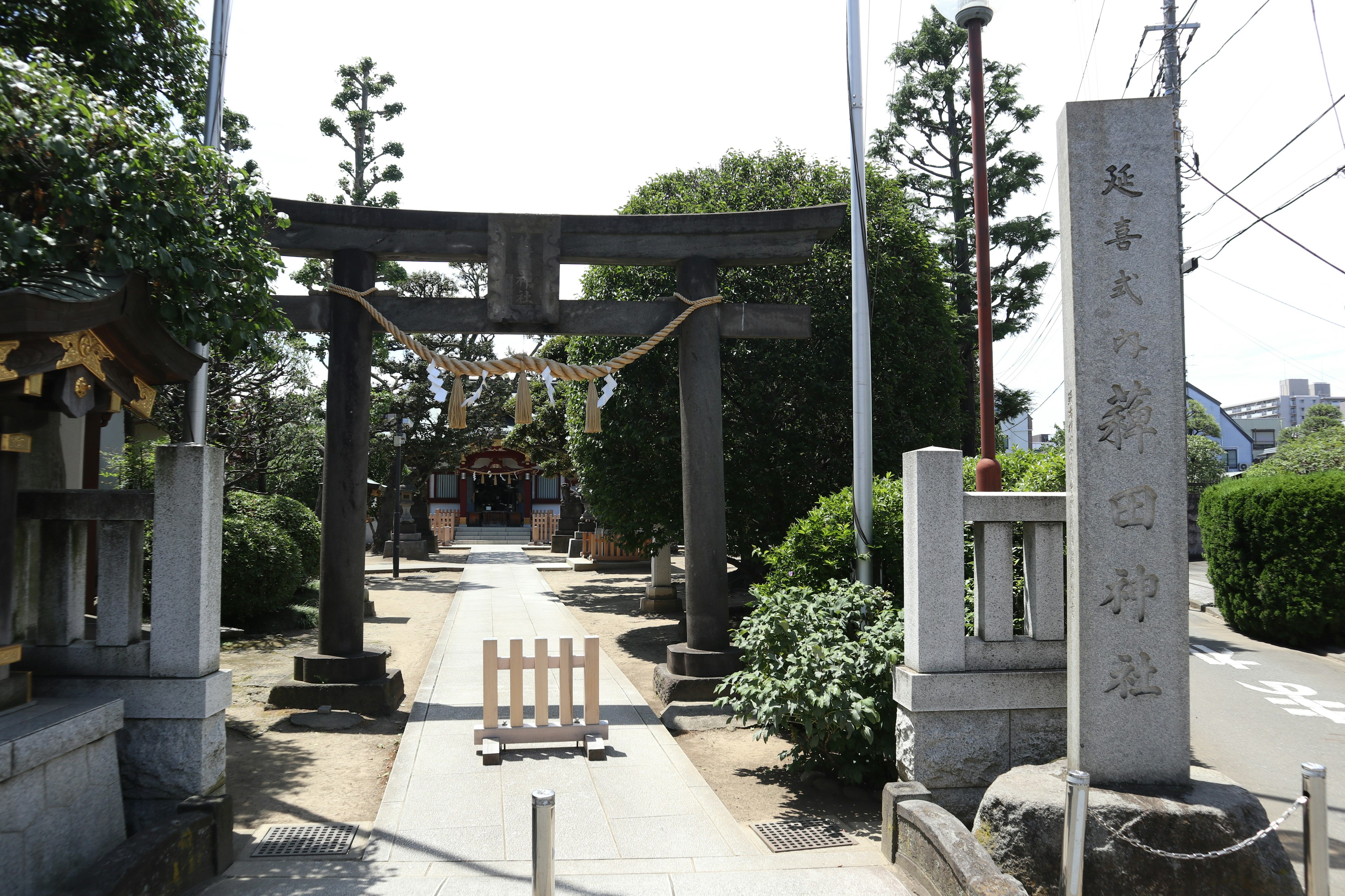 Cảnh cổng torii và lối vào đền thờ được bao quanh bởi cây cối và một tấm bia đá