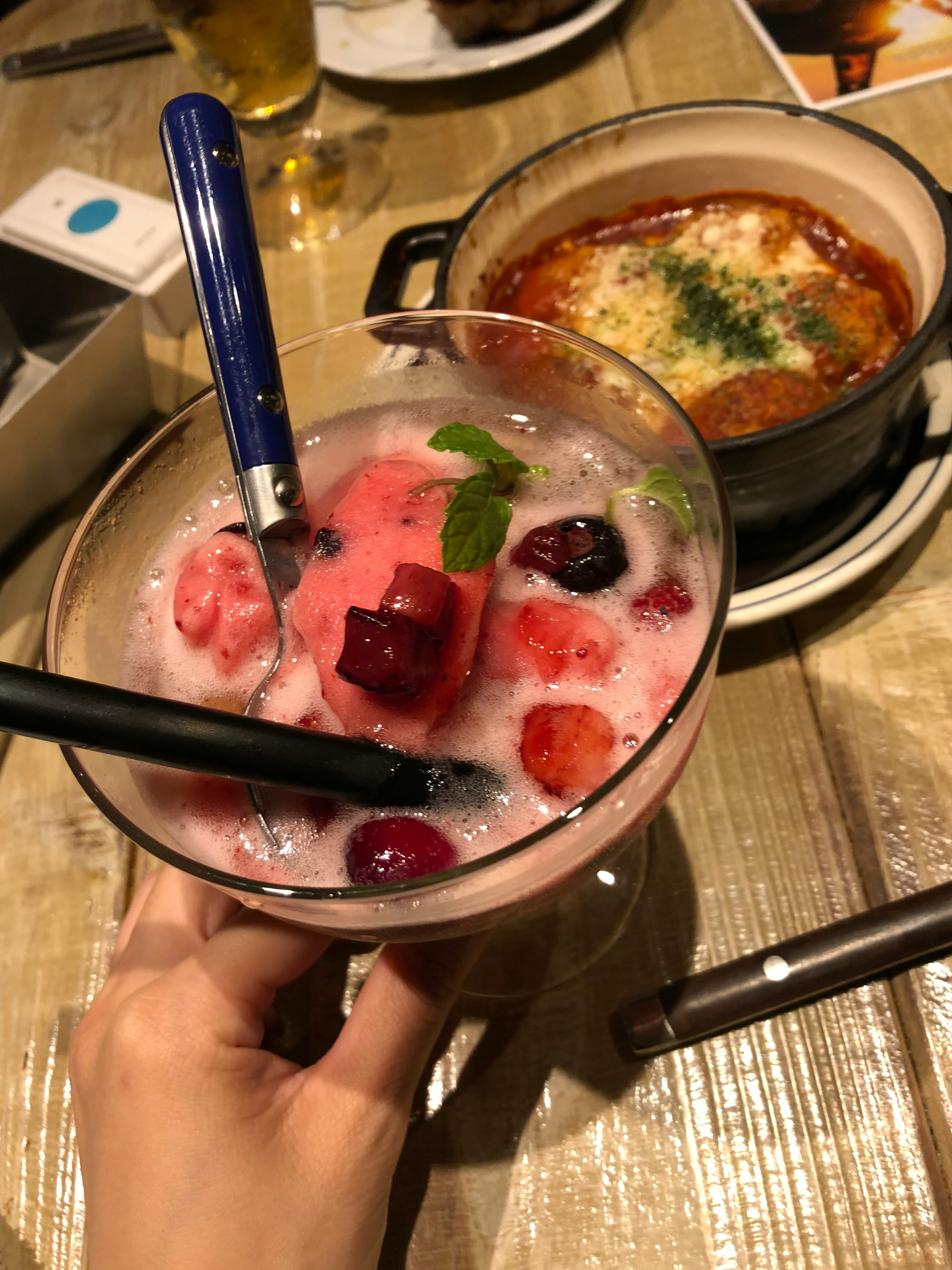 A colorful cocktail with berries in a glass held by a hand alongside a cheesy dish