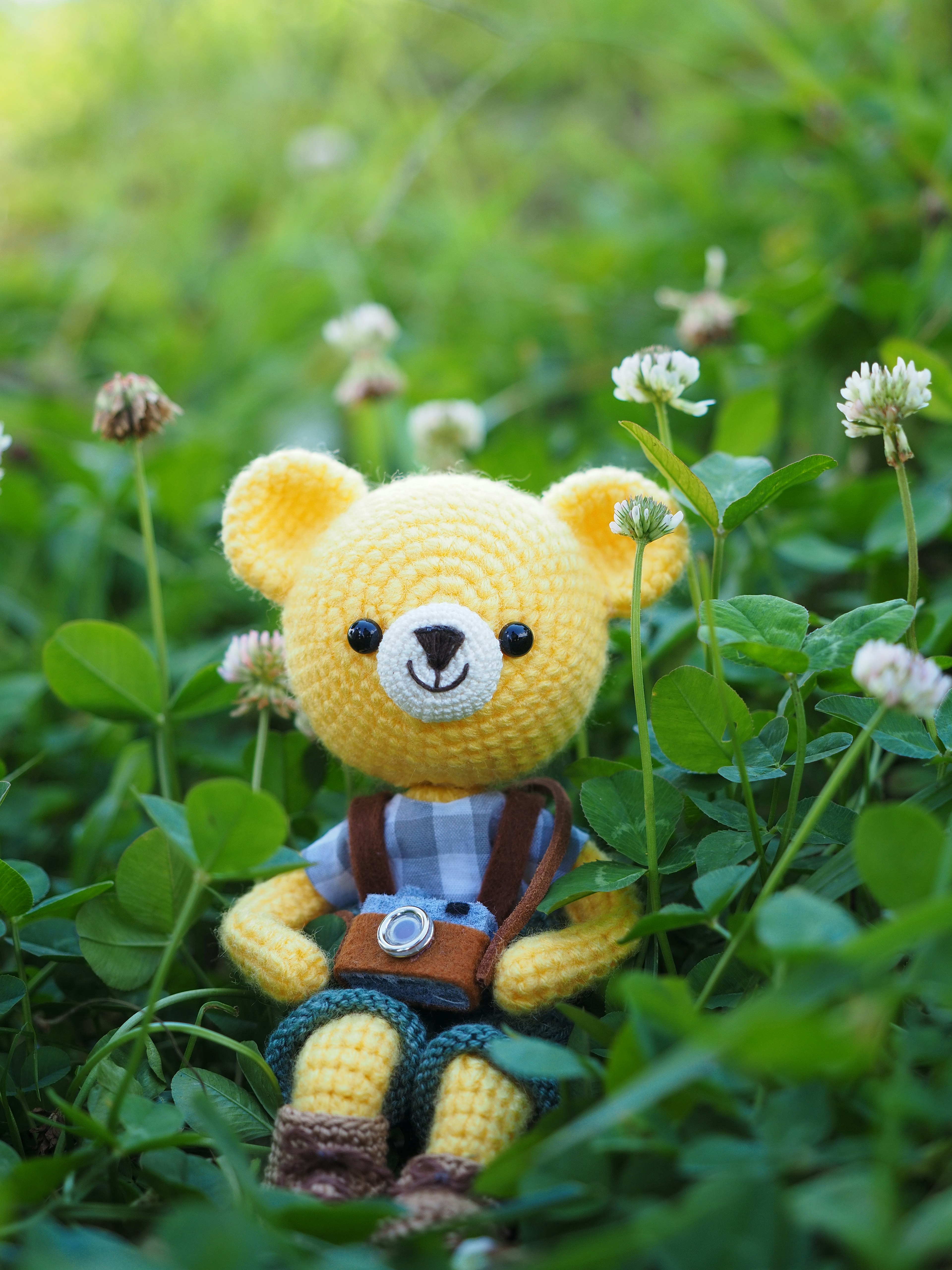 Crocheted yellow bear sitting among green clover and white flowers