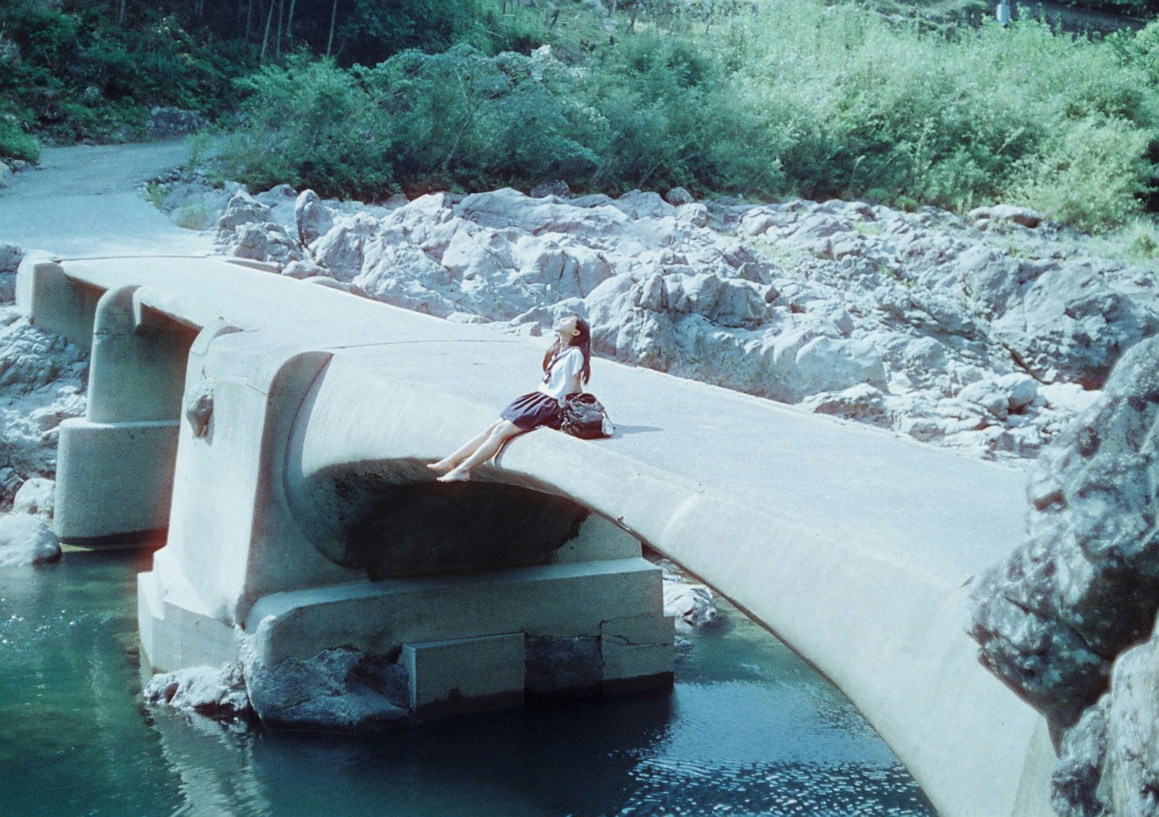 Femme assise sur un pont en béton entouré de verdure près d'un cours d'eau et de rochers