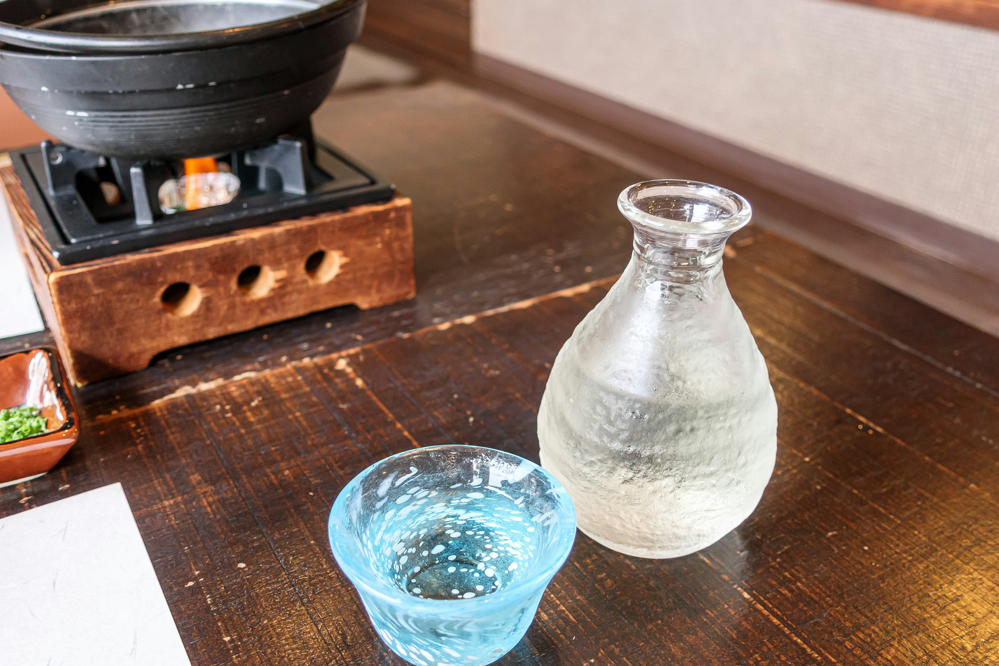 Una botella de sake transparente y una copa de sake azul sobre una mesa de madera