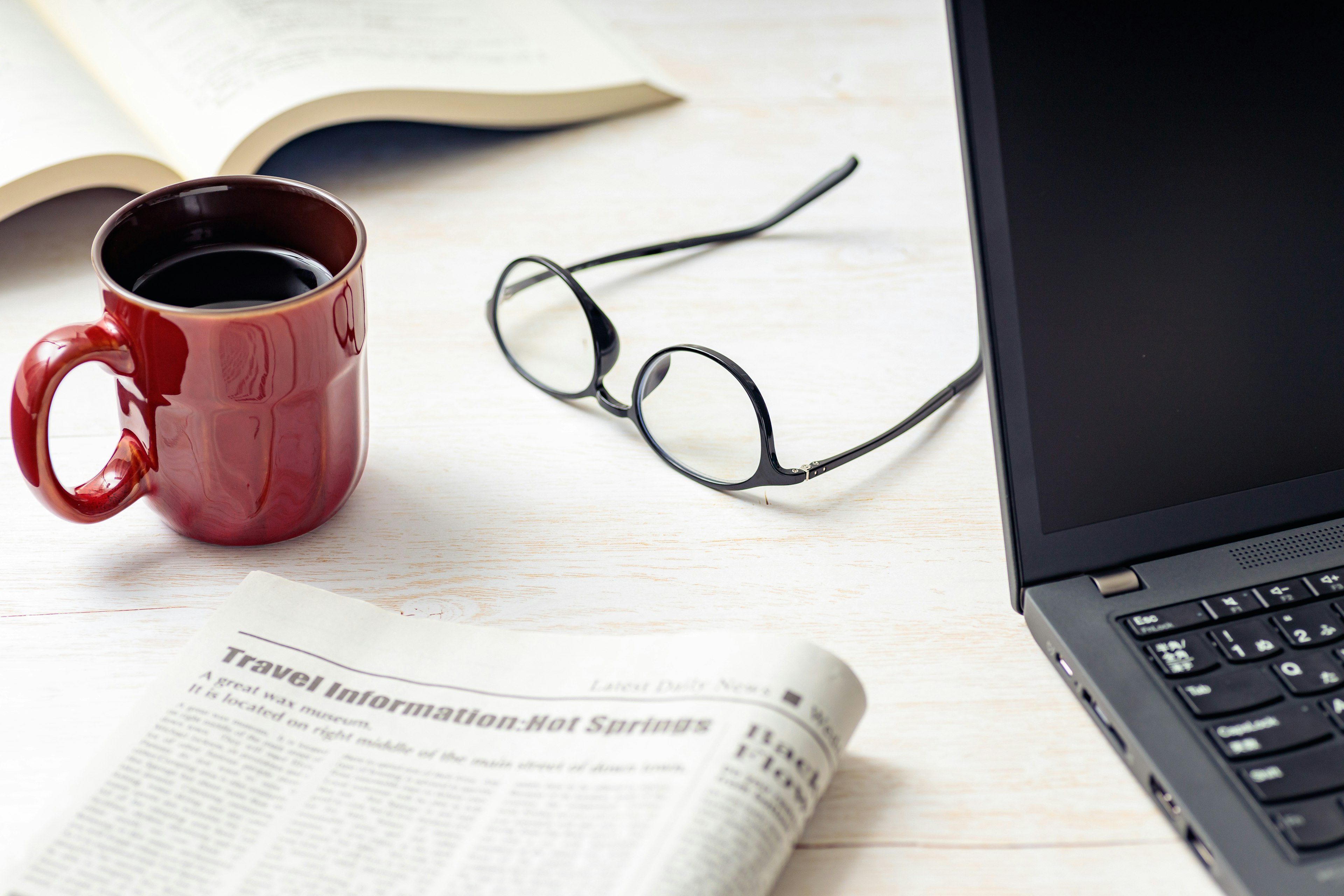 Taza roja sobre una mesa con gafas computadora portátil periódico y un libro abierto