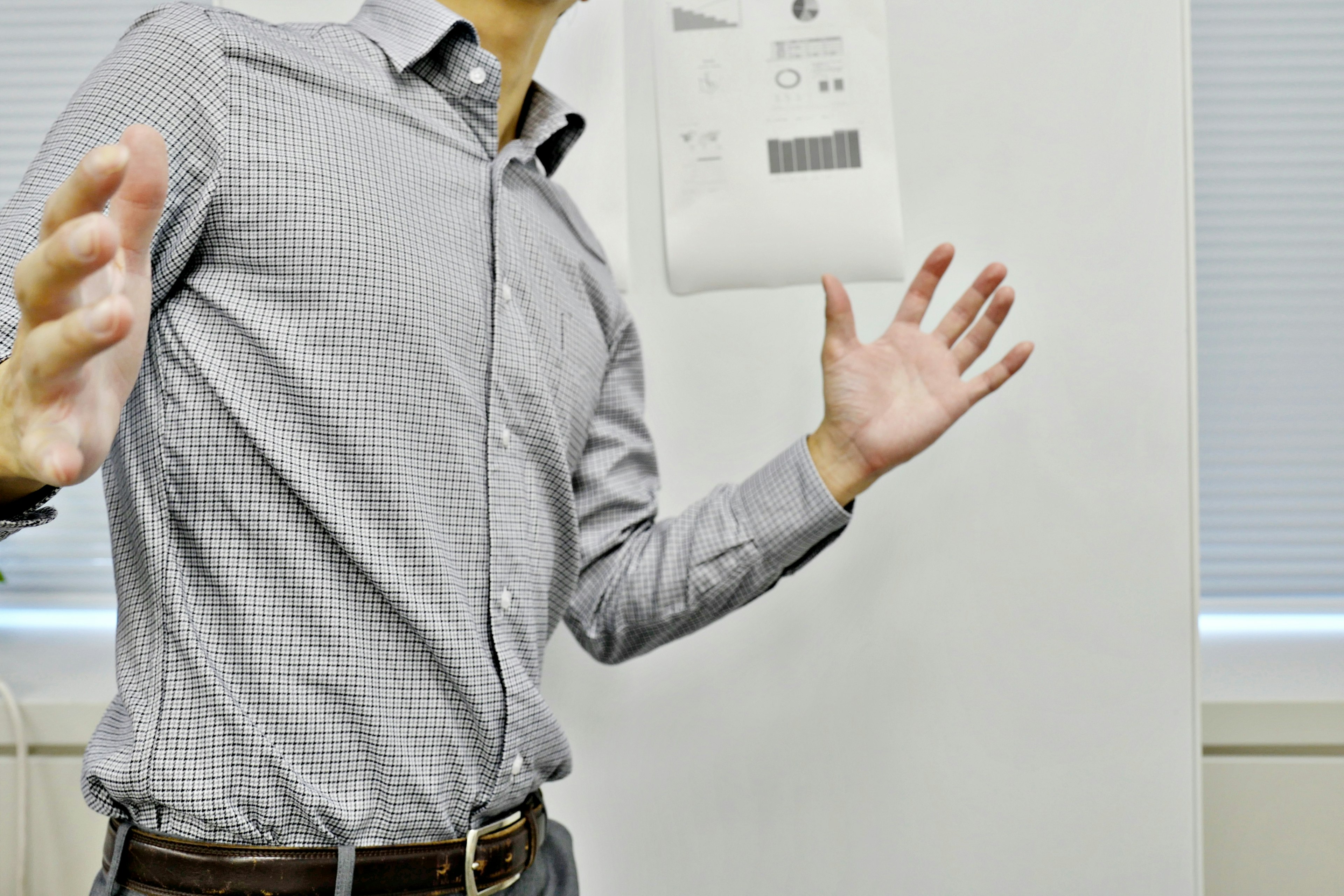 A man gesturing with hands in a checkered shirt and a white wall in the background