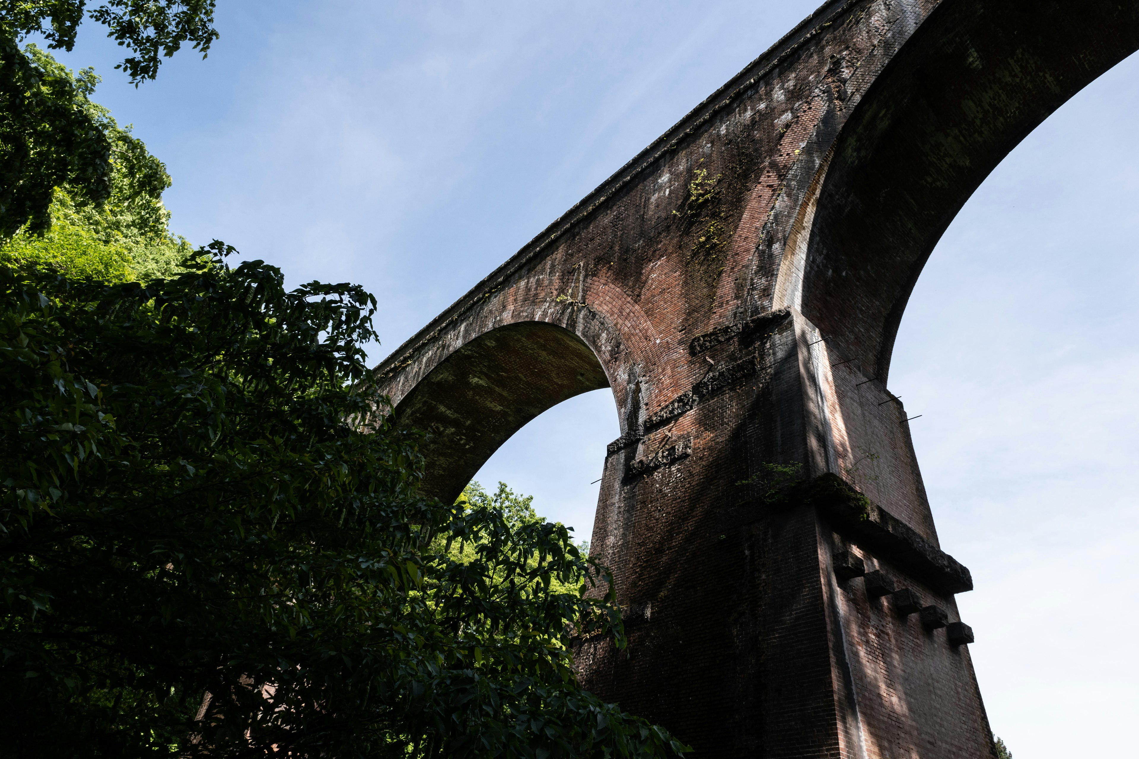 An old arch bridge towering under a blue sky surrounded by lush green trees