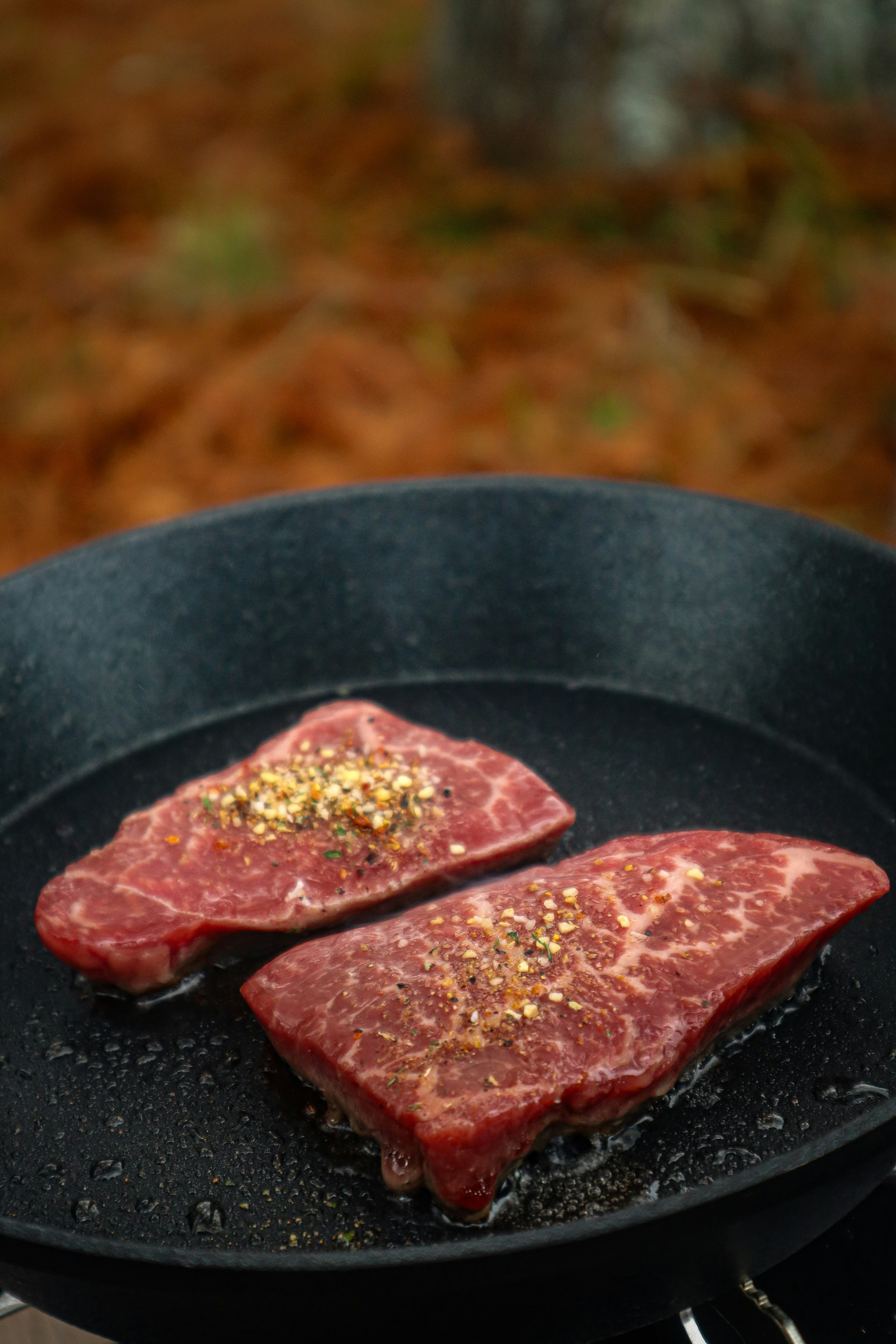 Two pieces of marbled meat with spices on a black frying pan