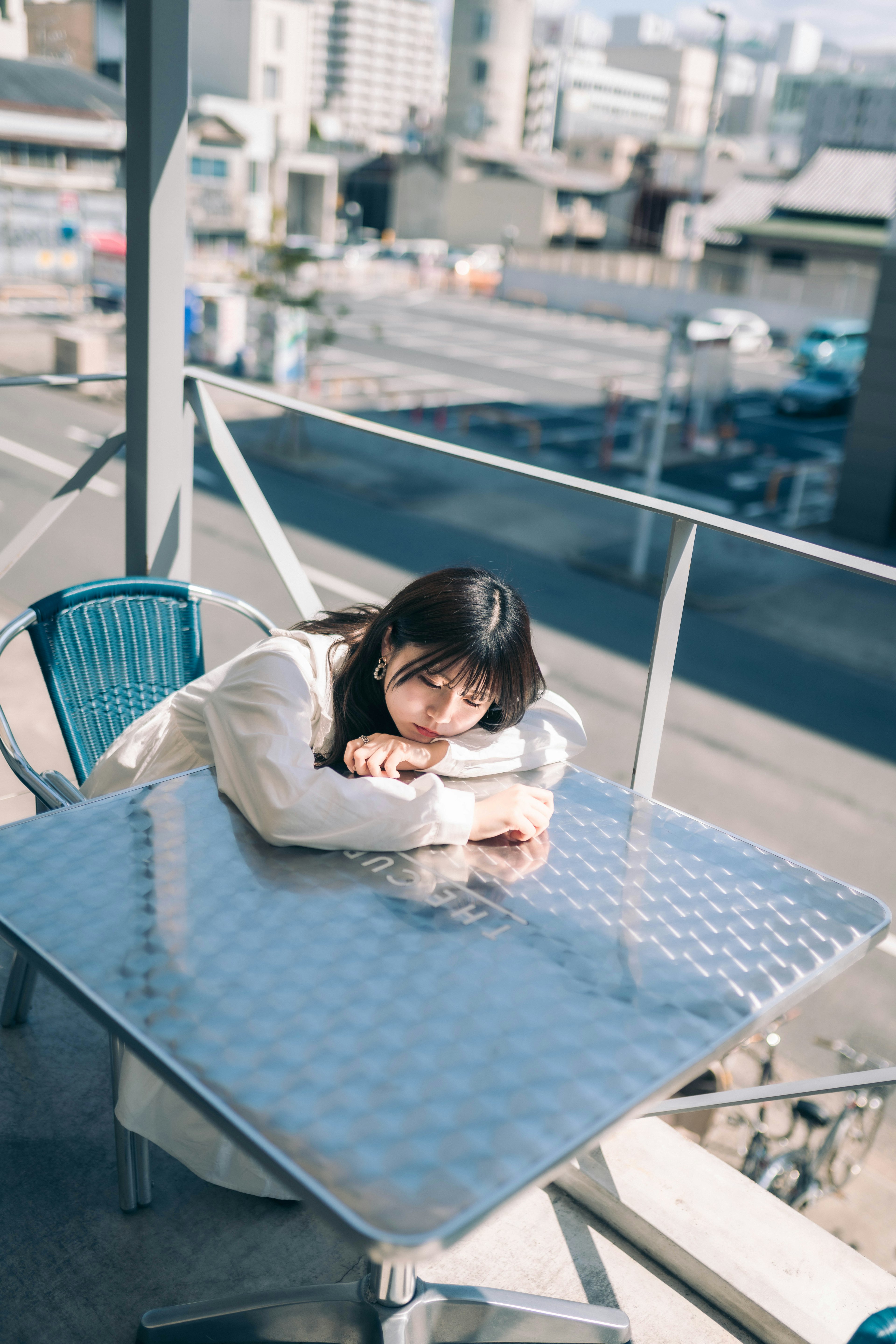 女性がテラスのテーブルに寄りかかっているシーン　背景には都市の風景が広がる