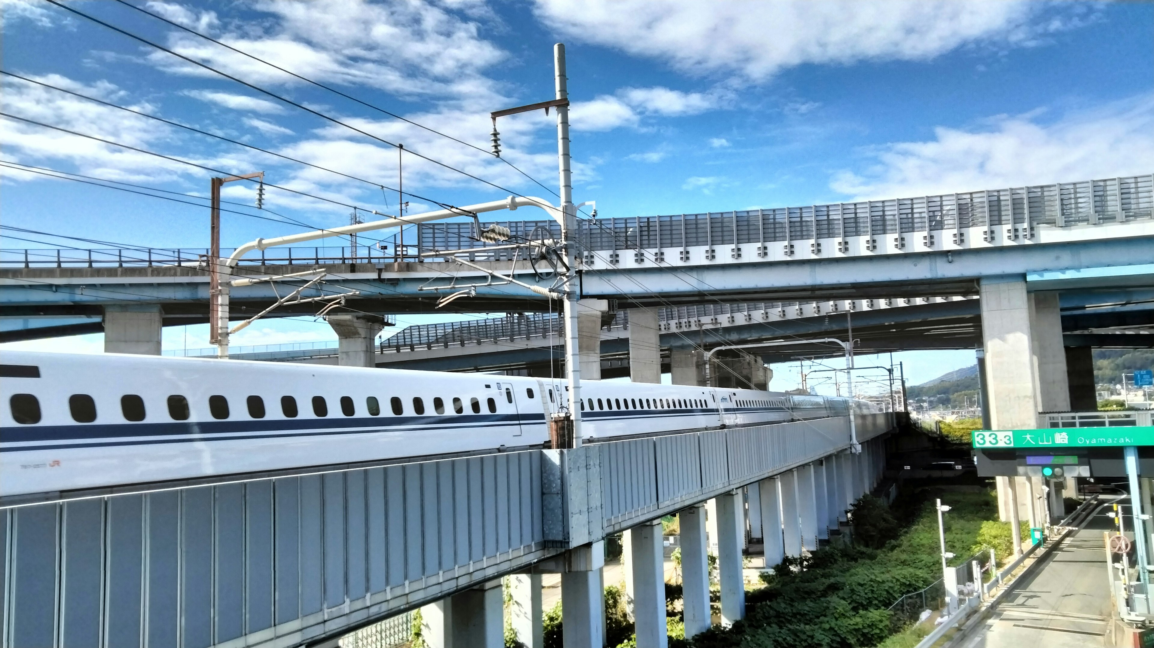 Shinkansen dan jalan tol bersilangan di bawah langit biru dengan awan