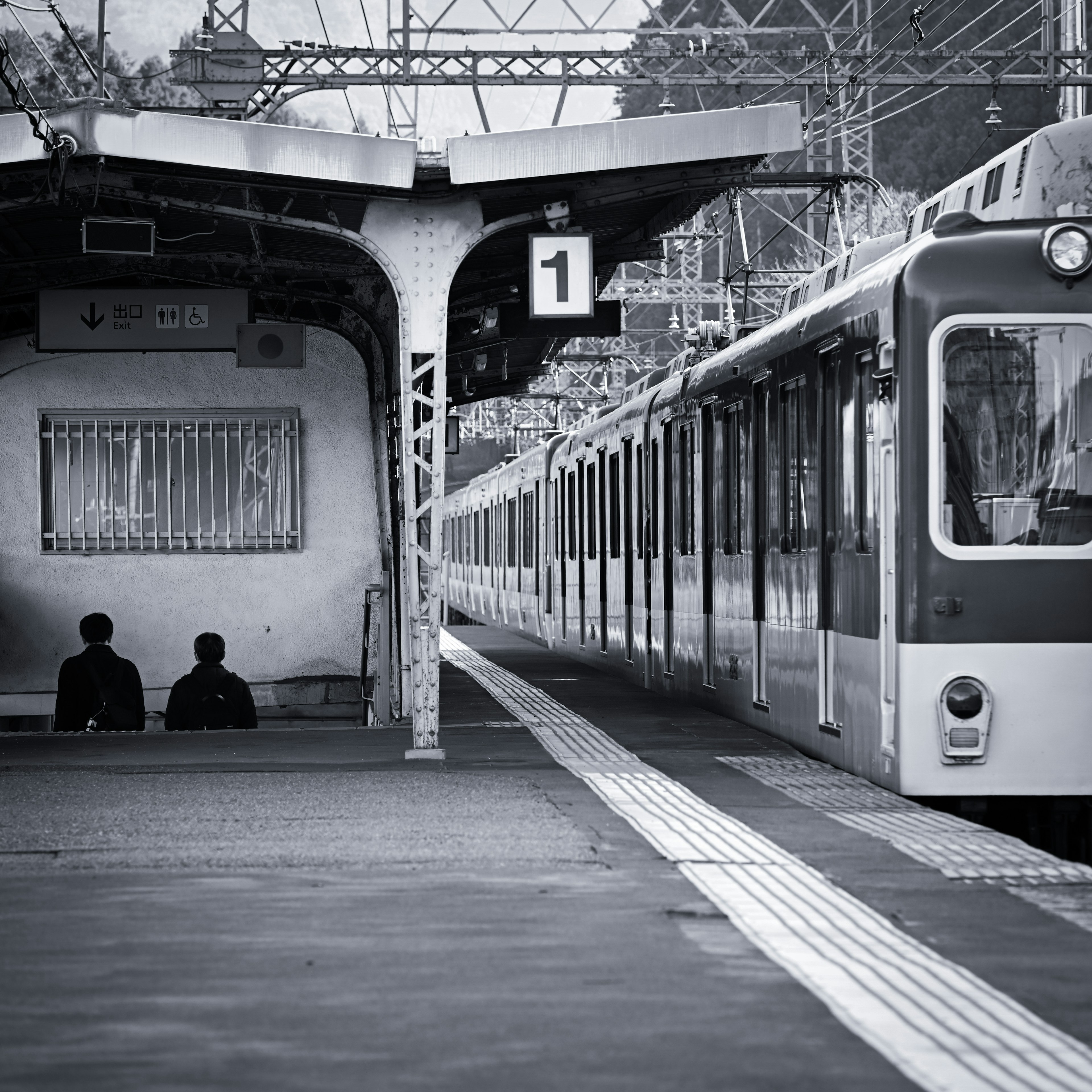 Imagen en blanco y negro de dos personas sentadas en una plataforma de tren con un tren detenido