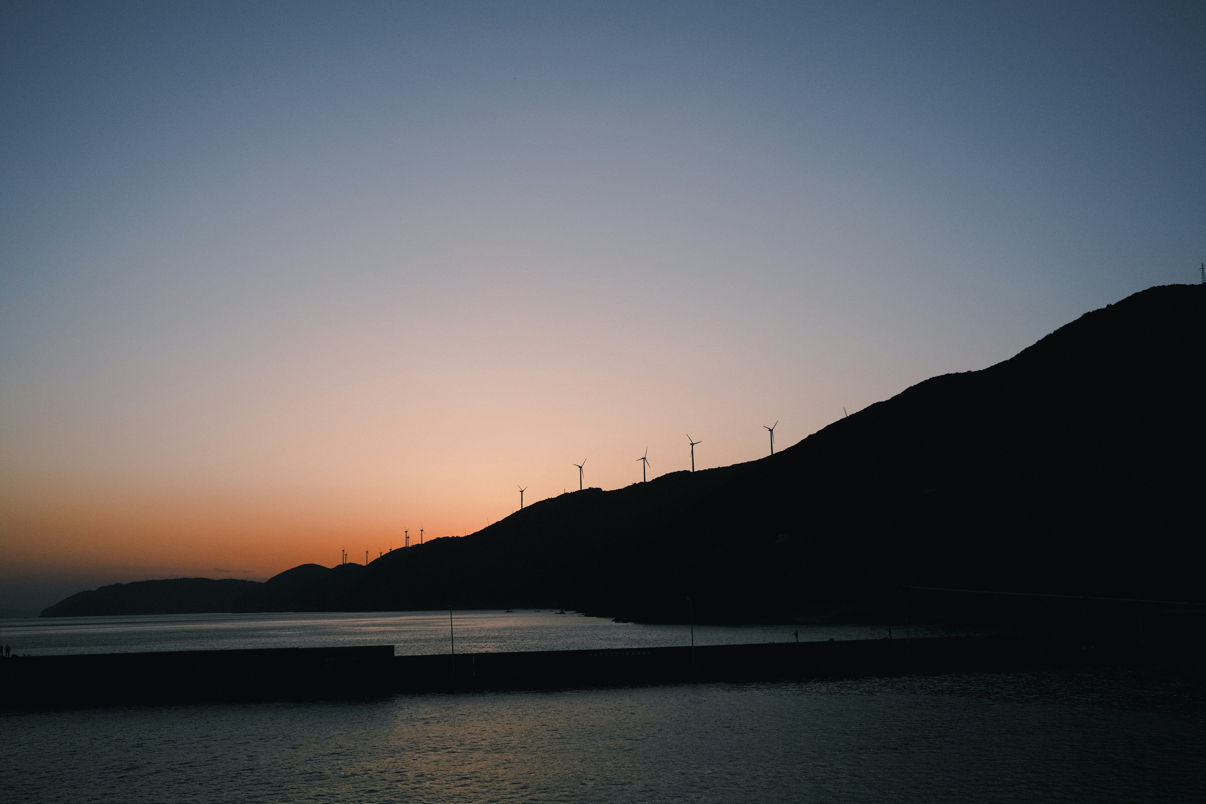 夕暮れ時の山のシルエットと風力発電機が見える海の風景