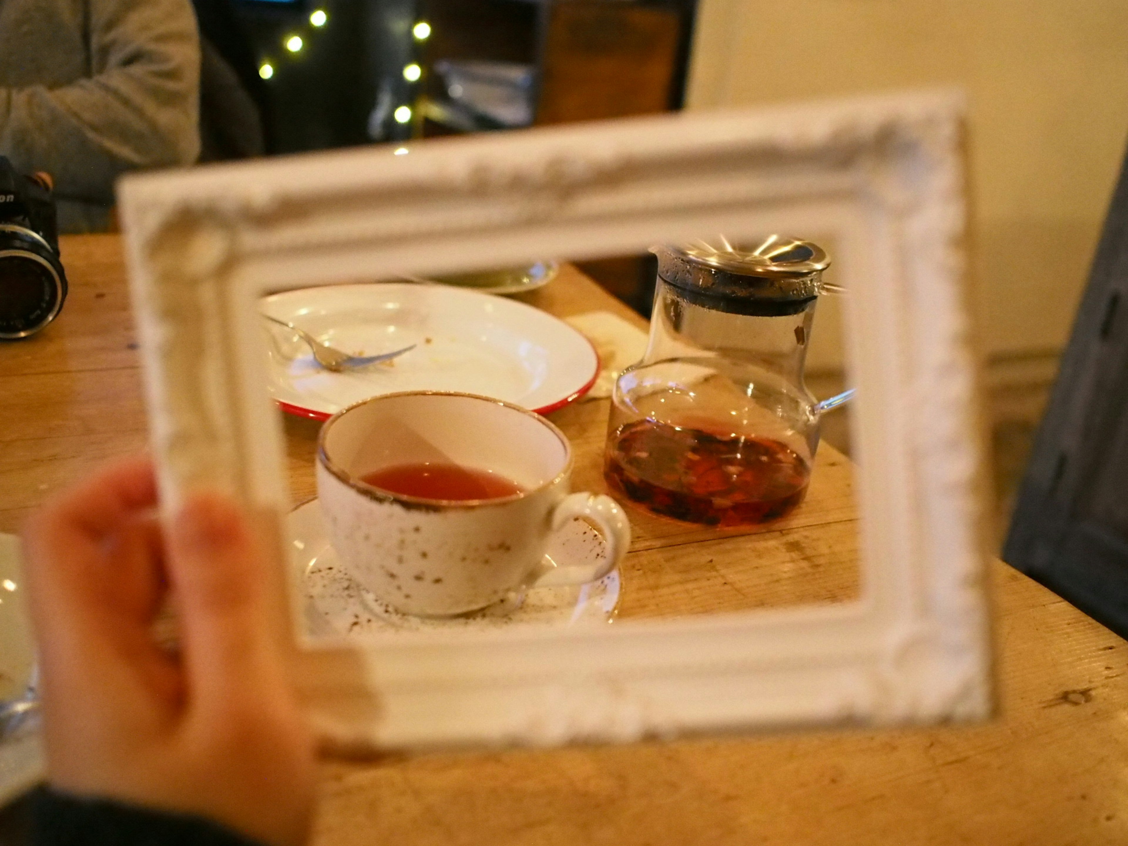 A hand holding a white frame around a cup of tea with a teapot and plate on the table