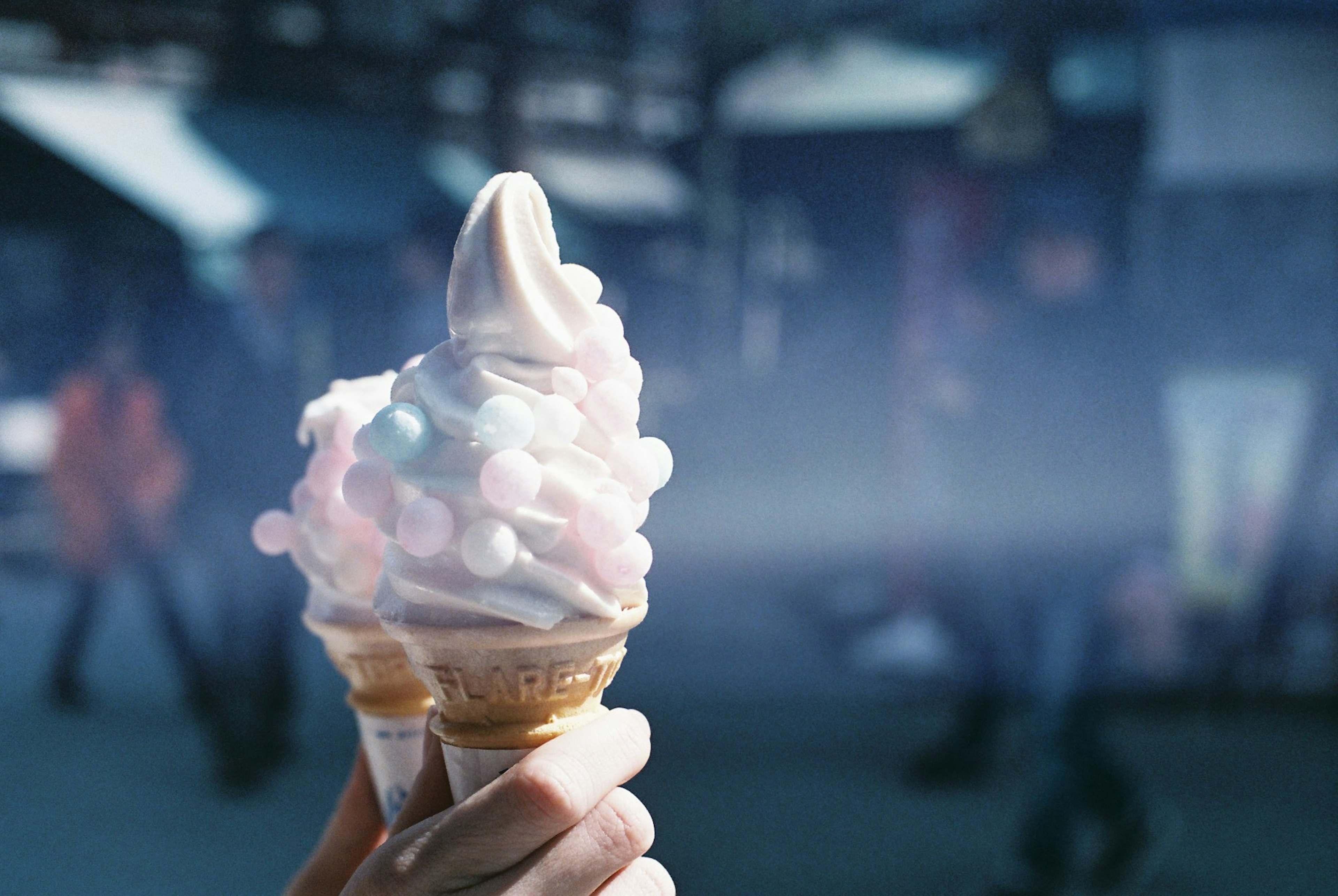 Hand, die einen Eisbecher mit Sahne und bunten Streuseln hält, vor einem verschwommenen Hintergrund von Menschen