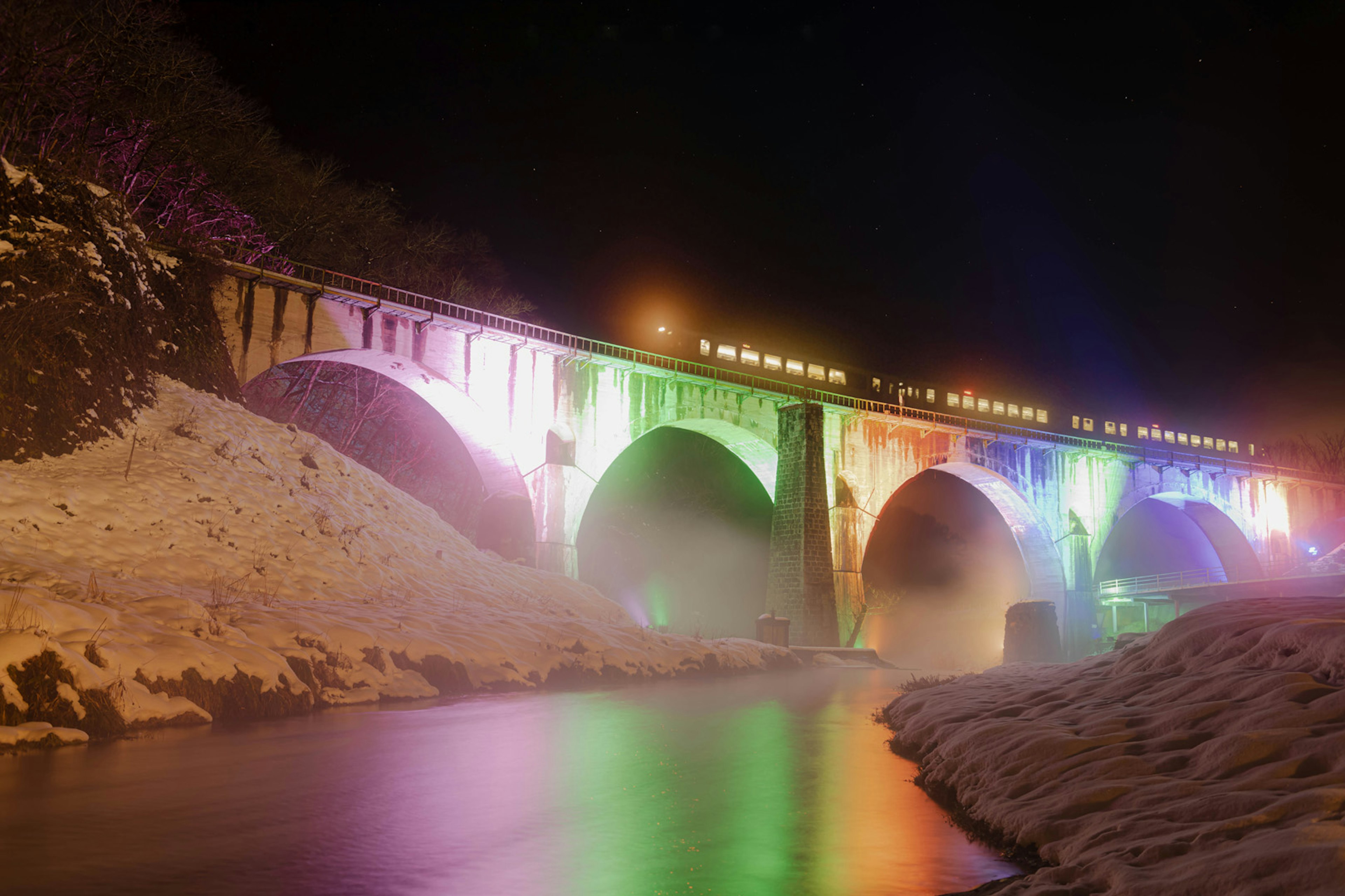 สะพานที่สว่างไสวในยามค่ำคืนสะท้อนสีบนแม่น้ำที่มีหิมะ