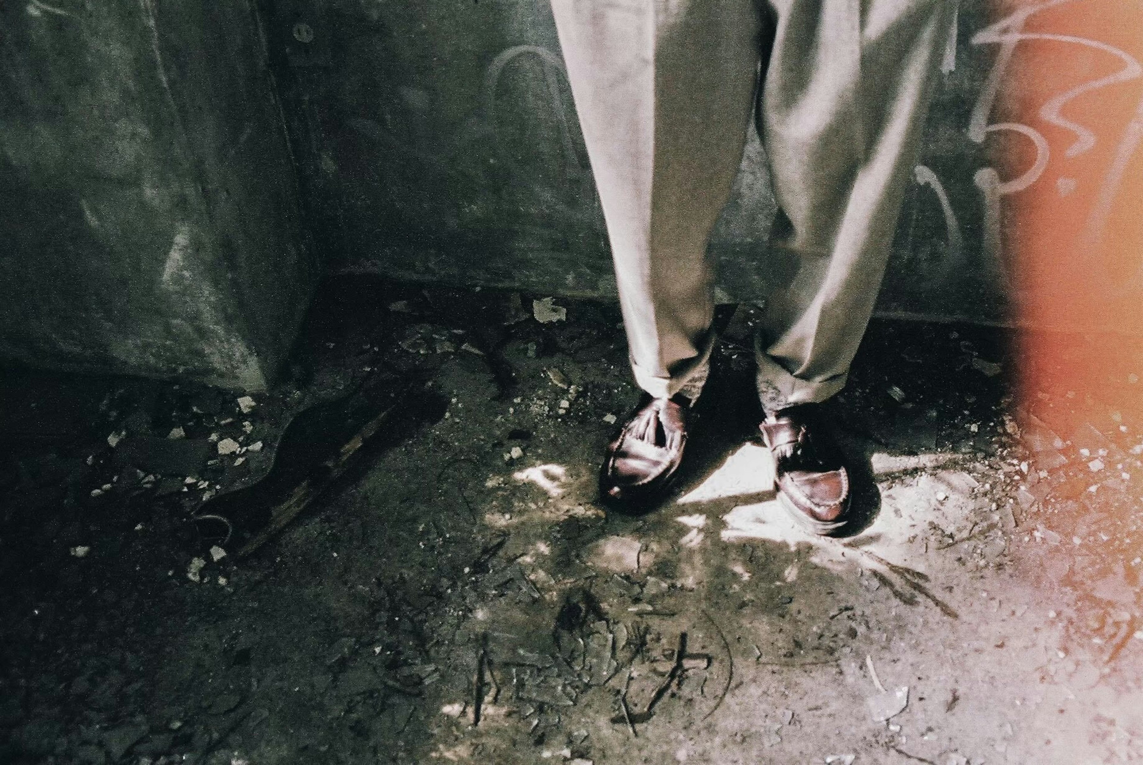 Feet of a person wearing light-colored pants standing in a dimly lit space with graffiti on the wall