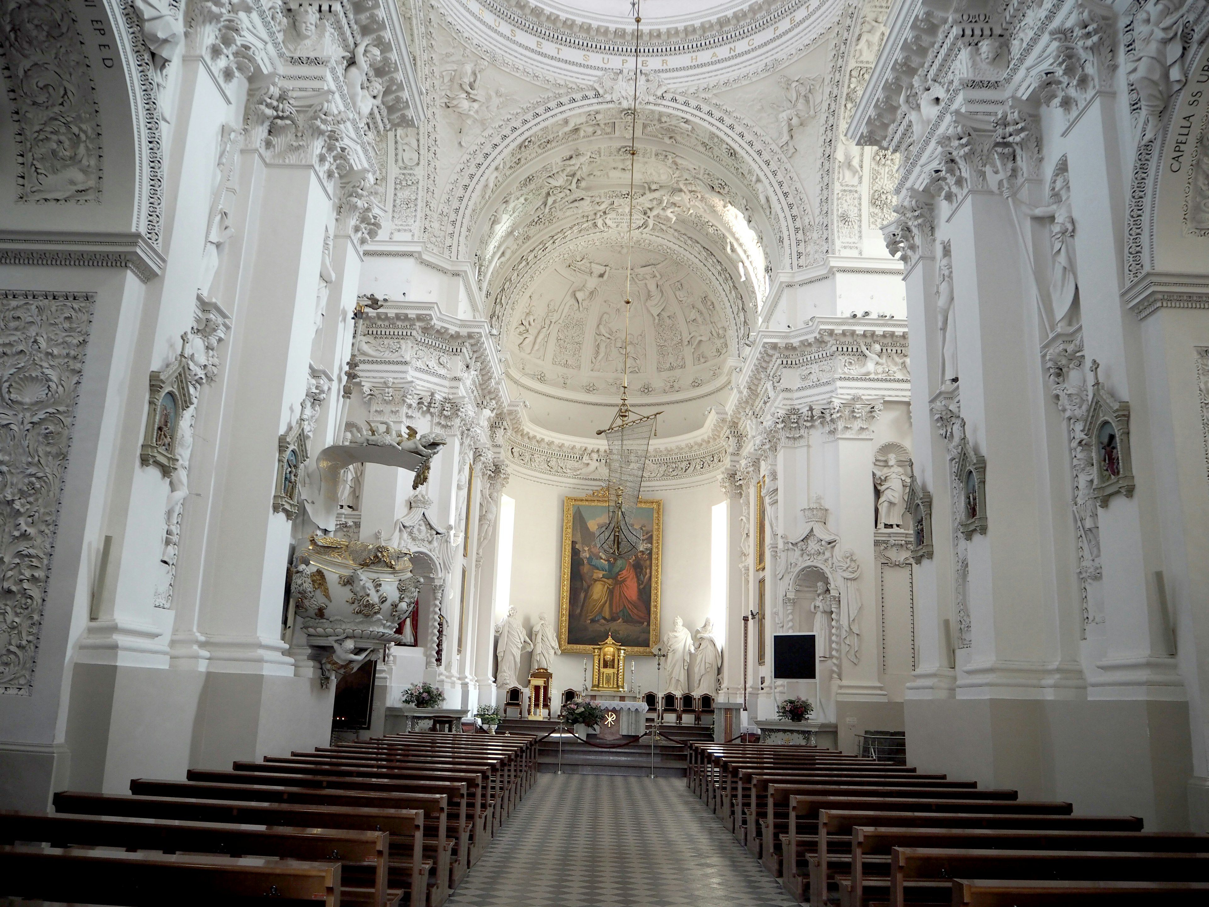 Interior of a church with white walls and ornate sculptures featuring an altar and a large painting