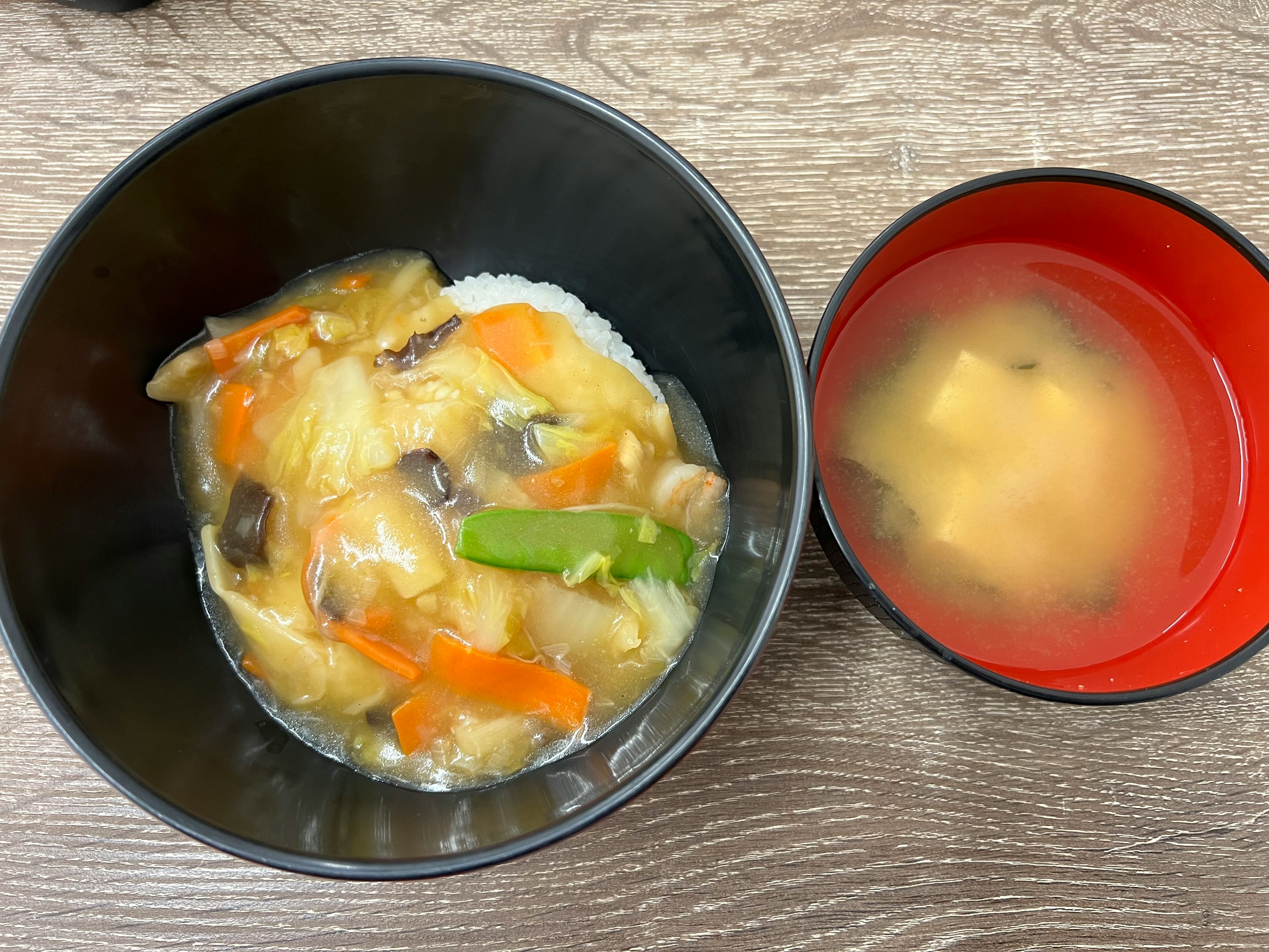 A bowl of rice topped with a savory chicken and vegetable sauce alongside a red bowl of miso soup