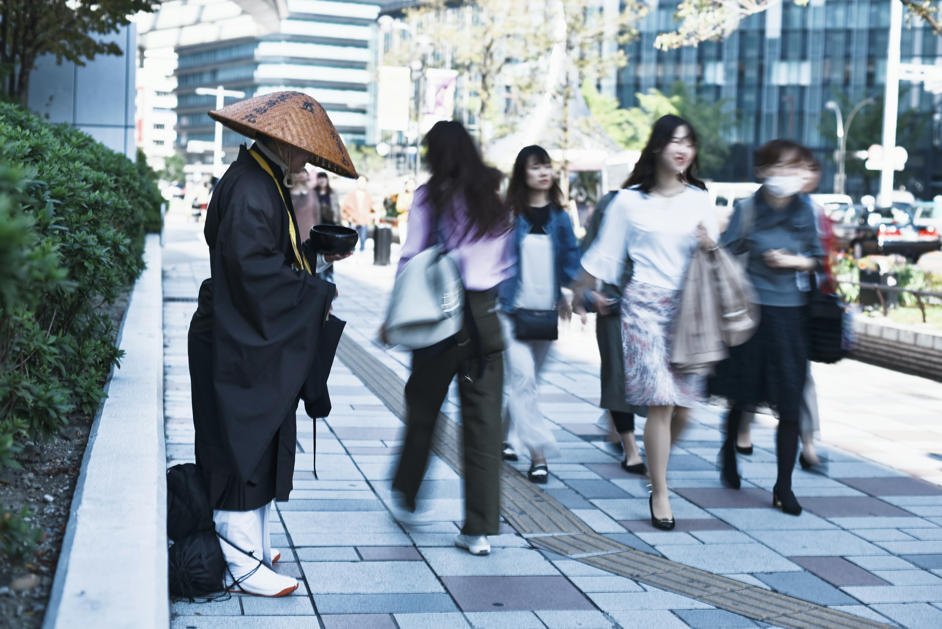 A monk sitting on the street surrounded by people walking in a modern urban setting