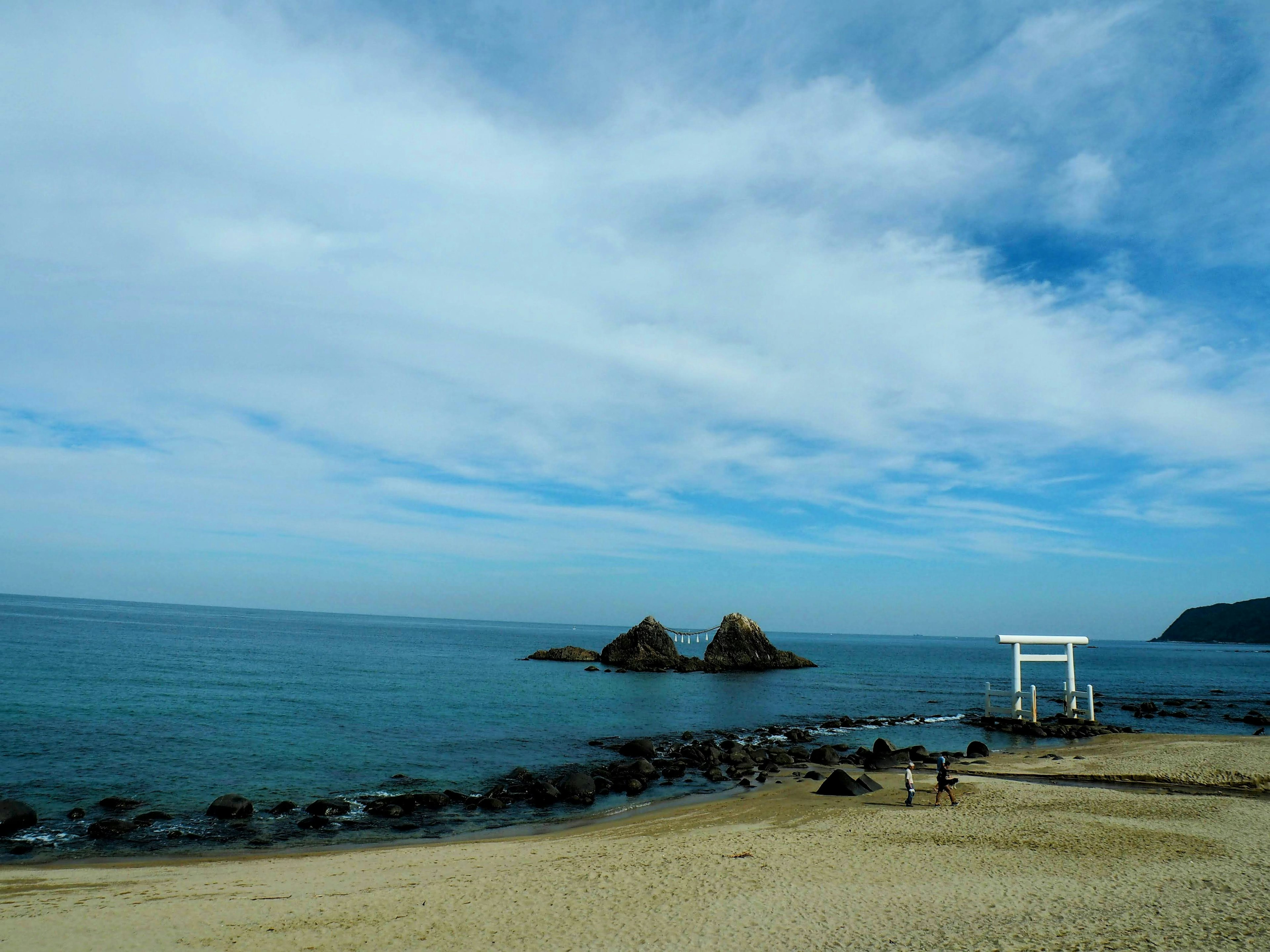 Pemandangan pantai berpasir dengan gerbang torii dan pulau berbatu di bawah langit biru