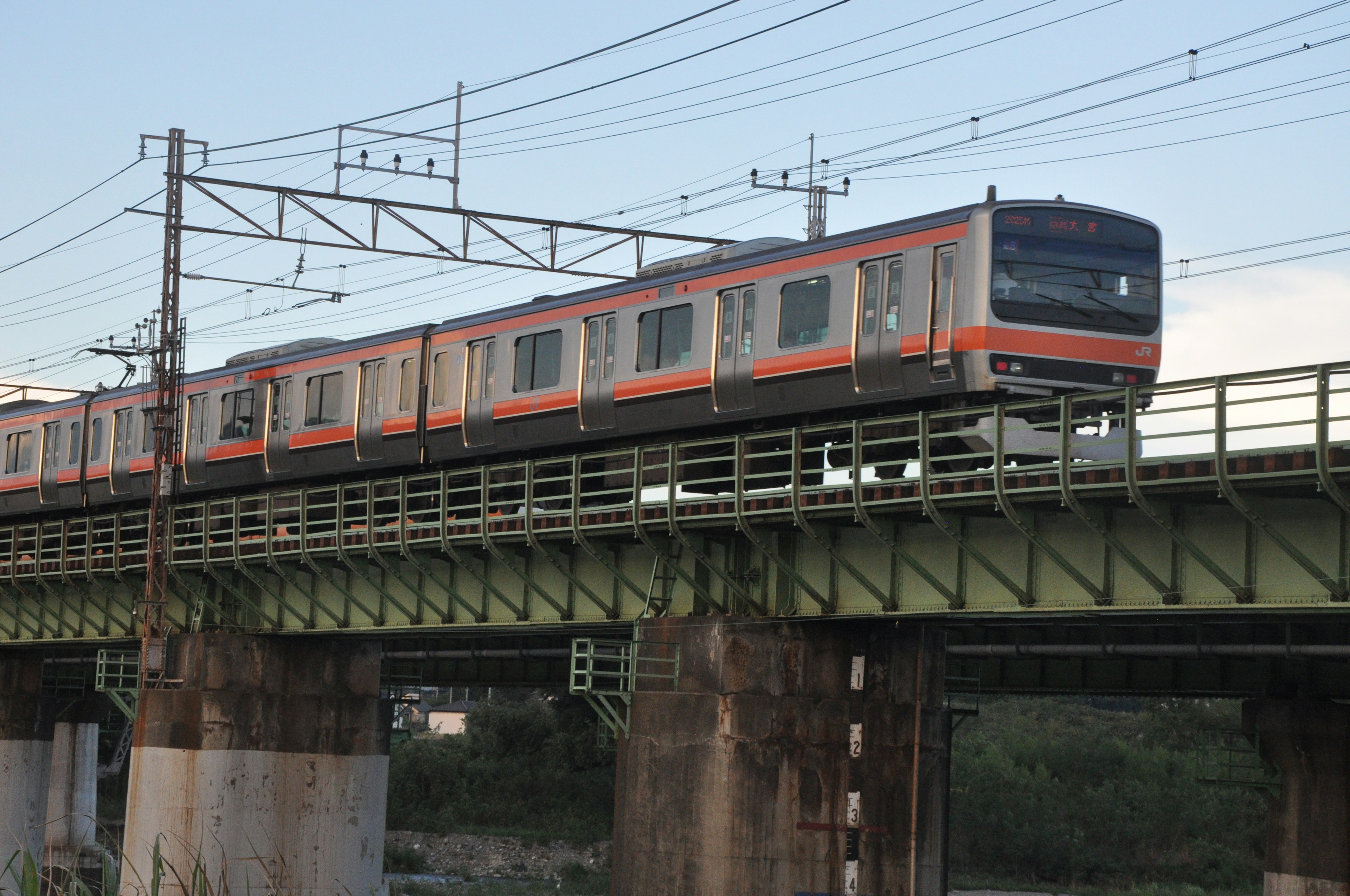 Train crossing a green bridge with overhead wires