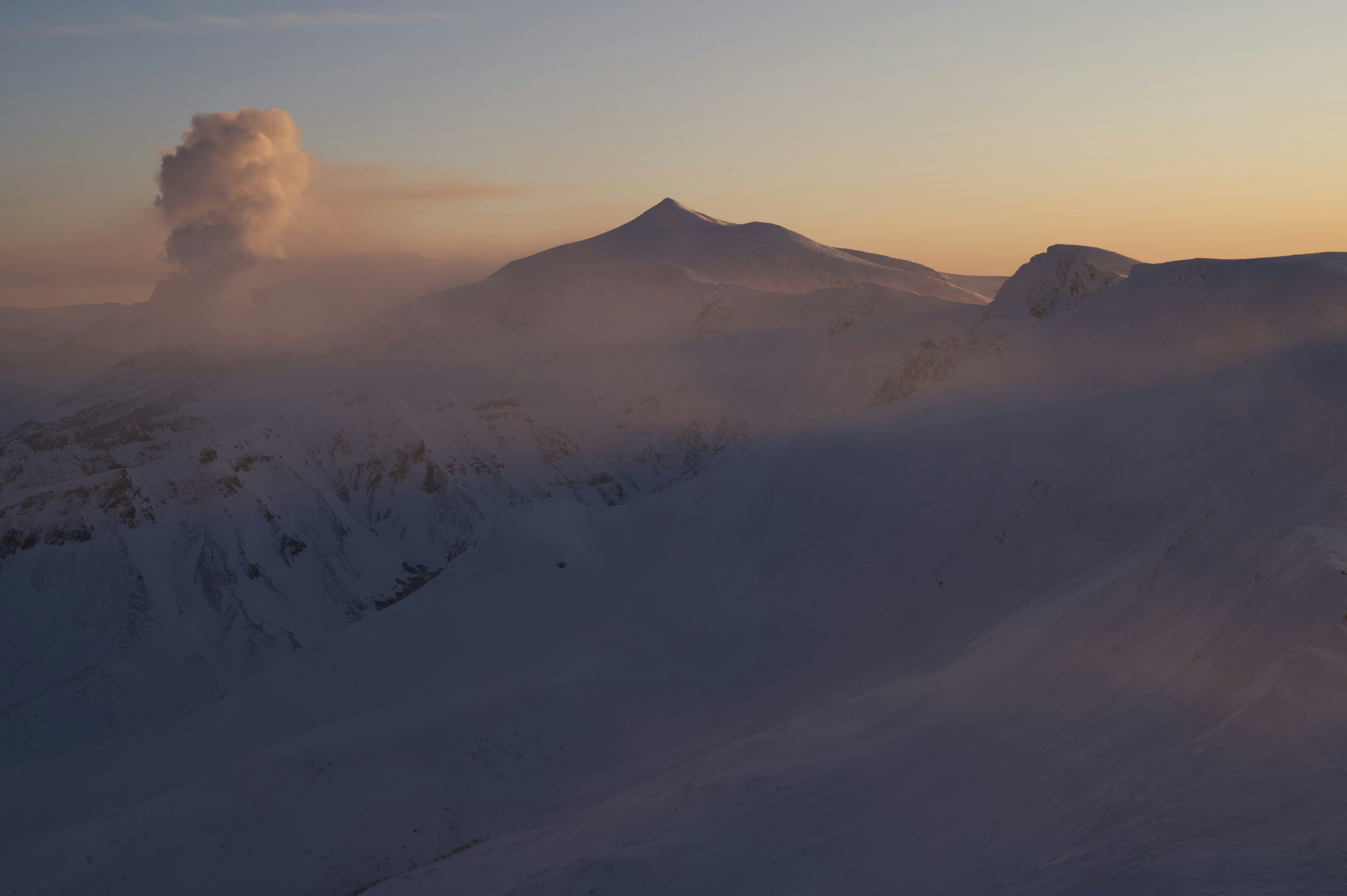 Snow-covered mountains with a volcano emitting smoke in the background