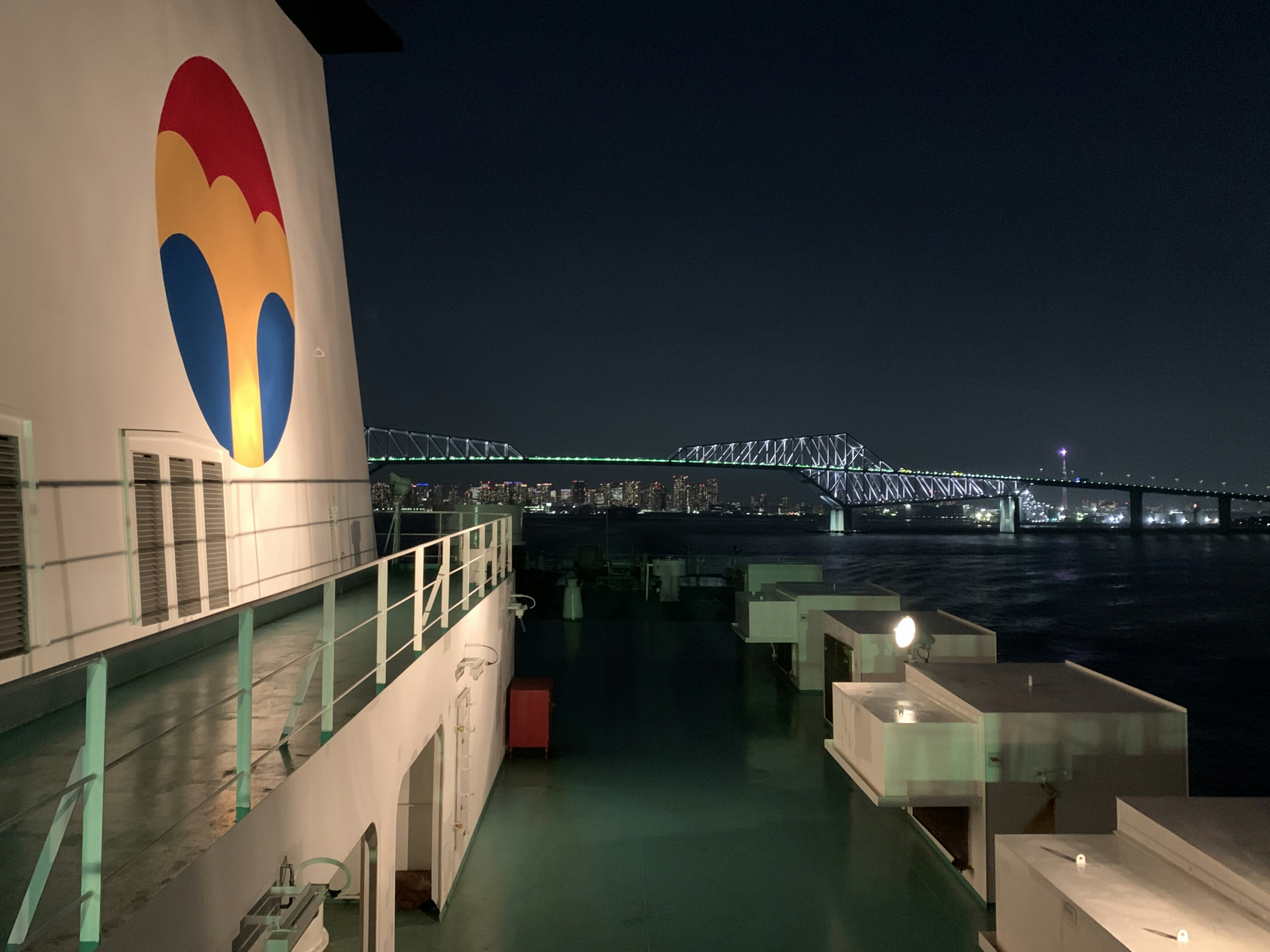 Side view of a ship at night with a colorful logo and a bridge in the background