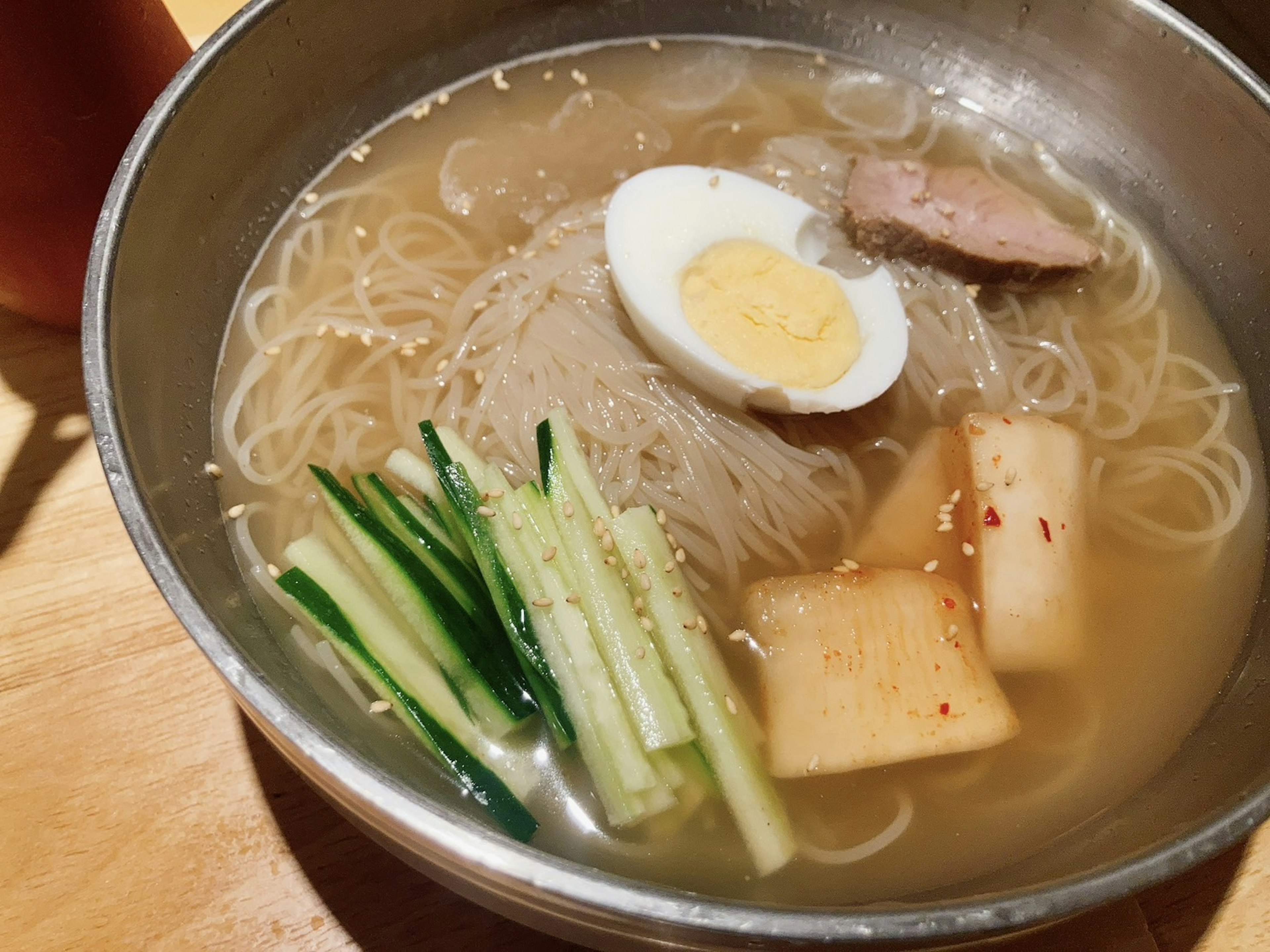 Bowl of clear broth with thin noodles topped with half a boiled egg sliced meat cucumber and pickled radish