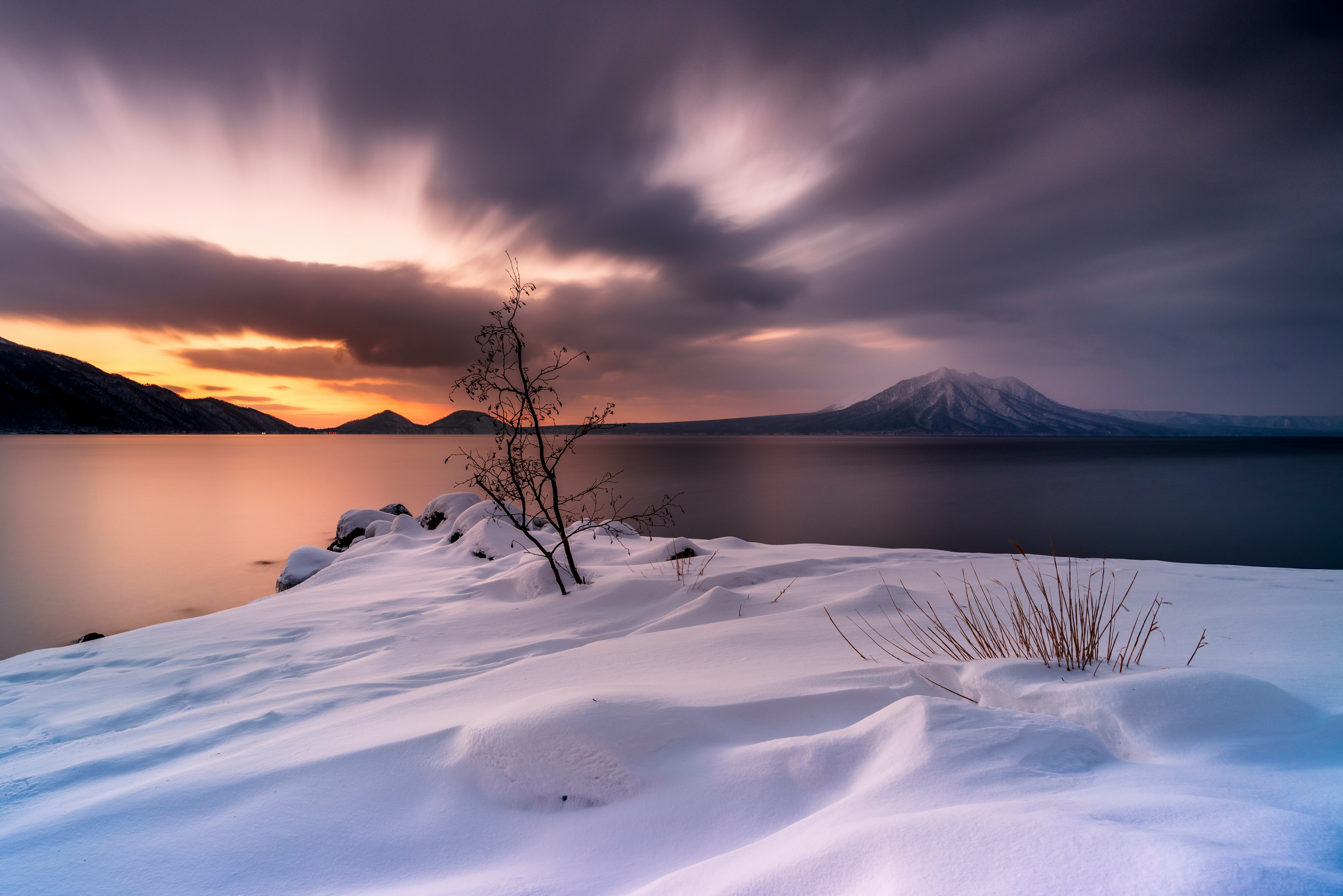 雪に覆われた湖の岸辺と背後の山々を映した夕暮れの風景