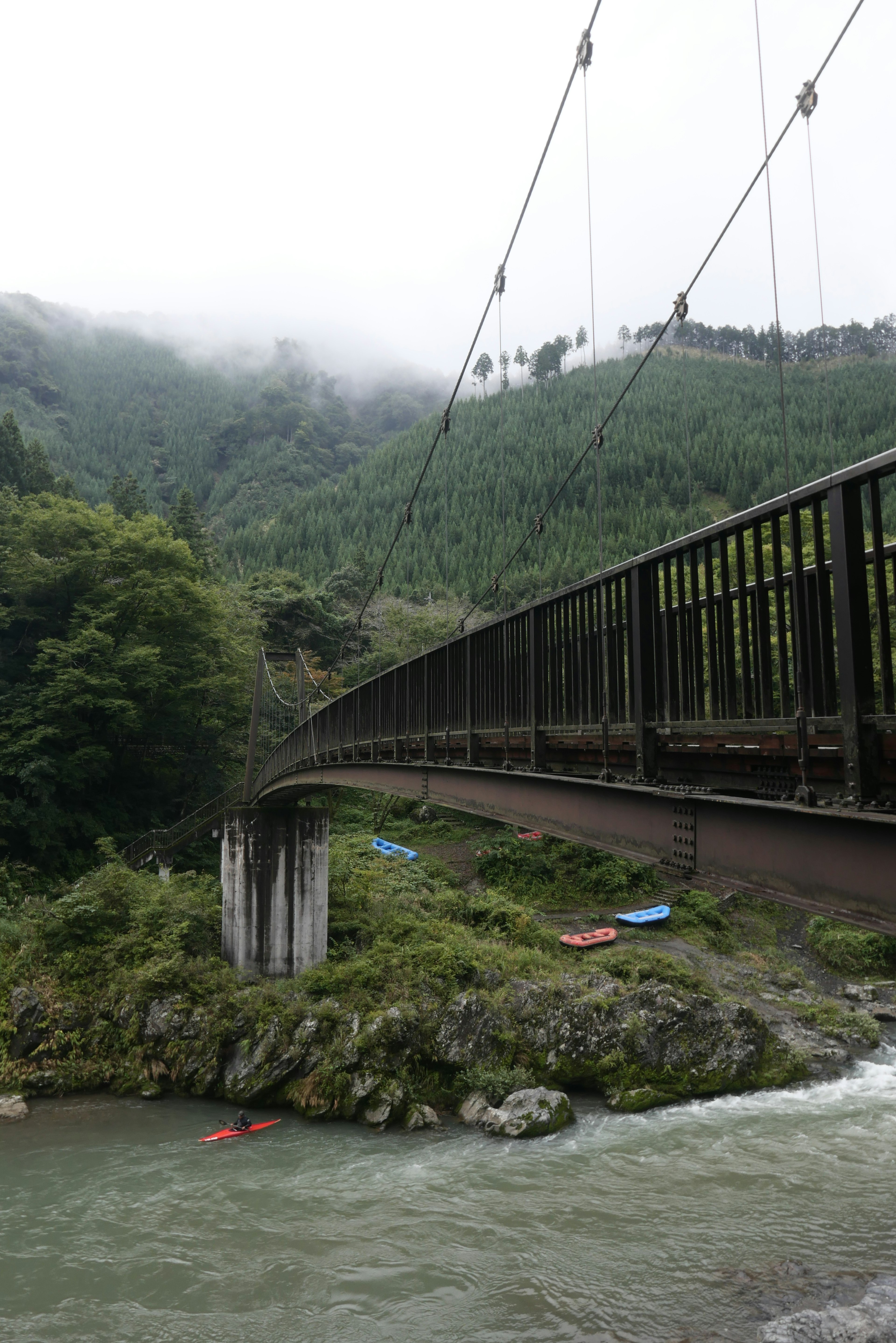 Hängebrücke über einen Fluss, umgeben von nebligen Bergen und Grünflächen