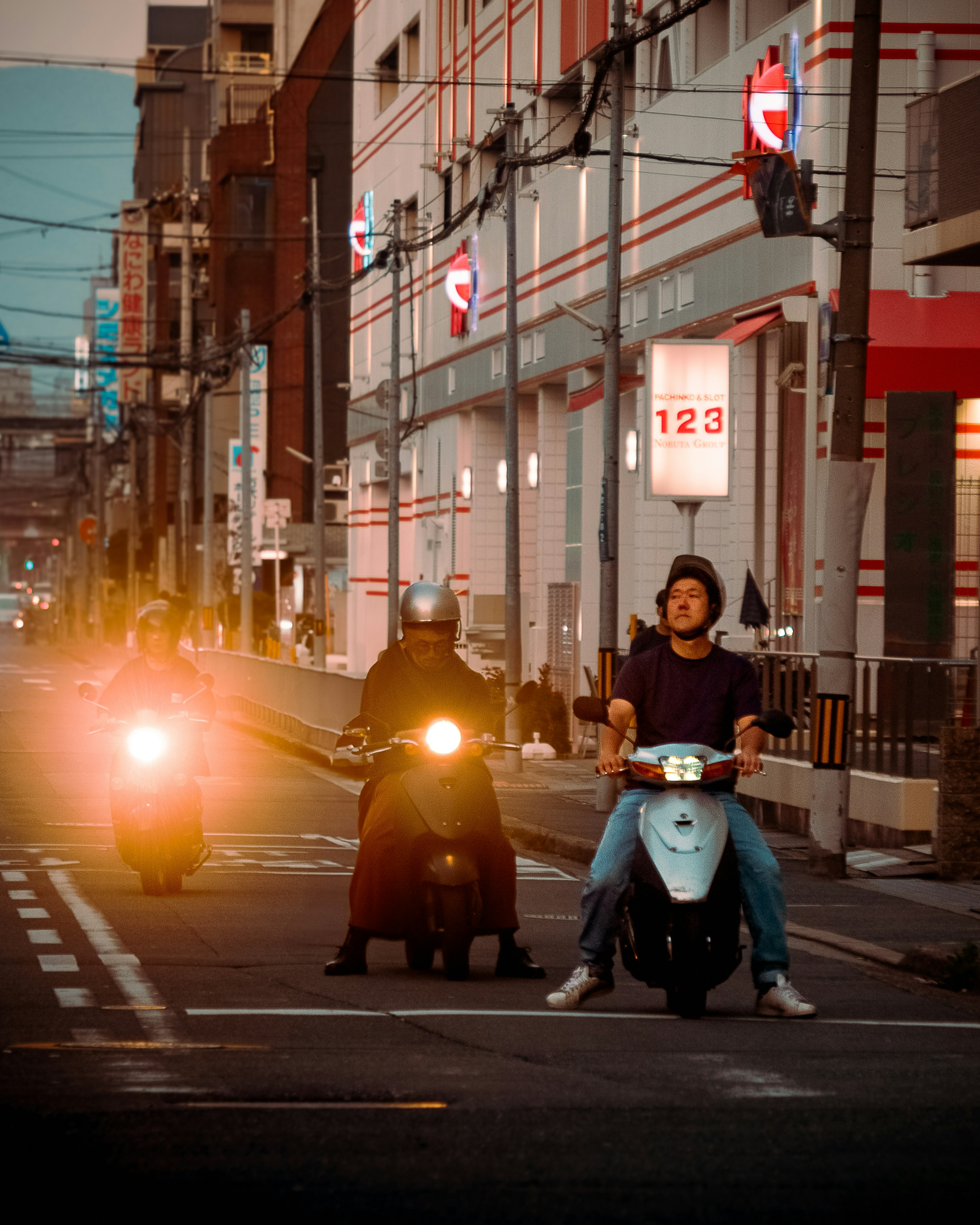 Three riders on scooters in a city street at dusk