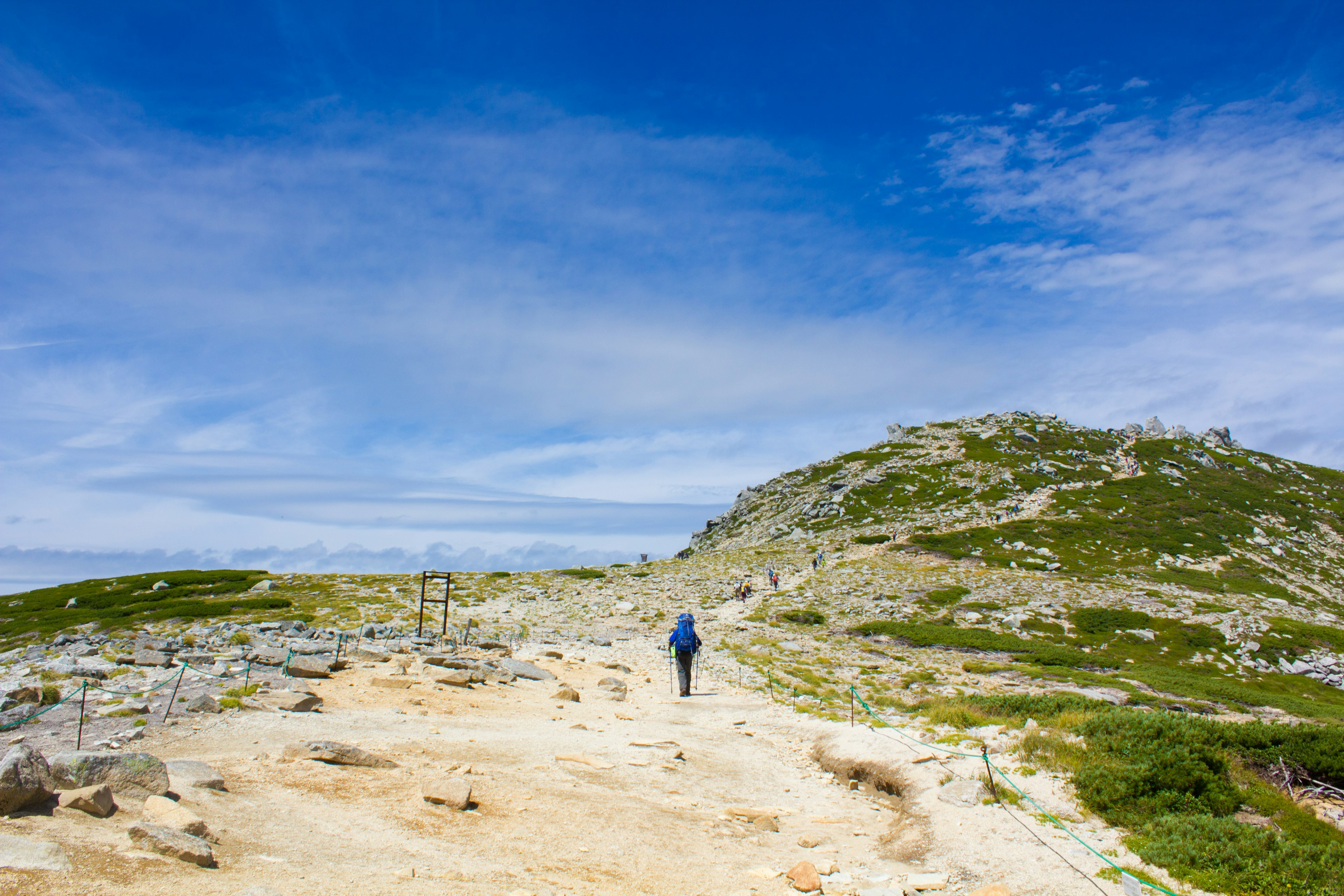 Una persona caminando por un sendero de montaña bajo un cielo azul y colinas verdes