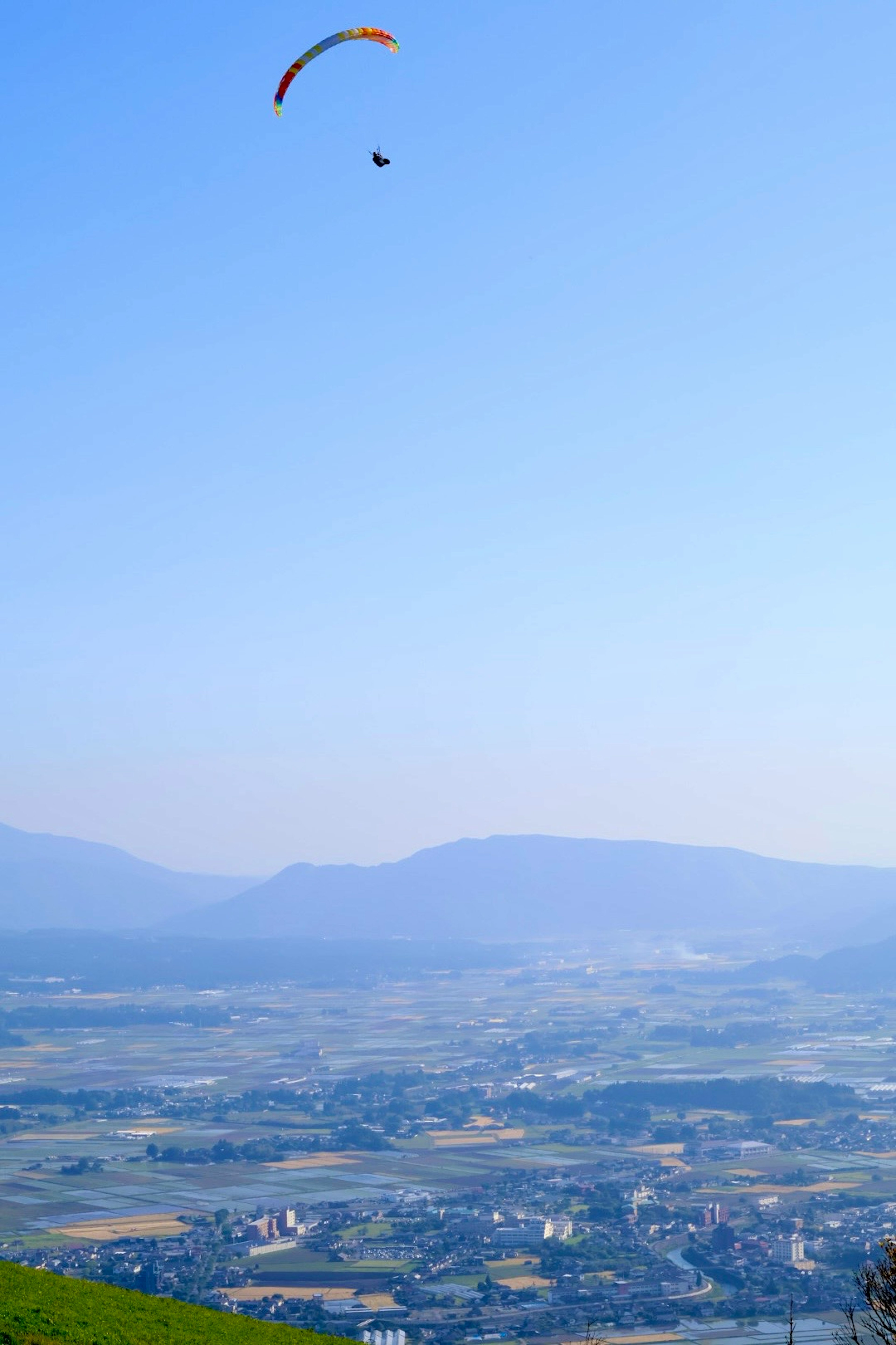 在蓝天之下，一名滑翔伞运动员在飞行，背景是山脉和山谷