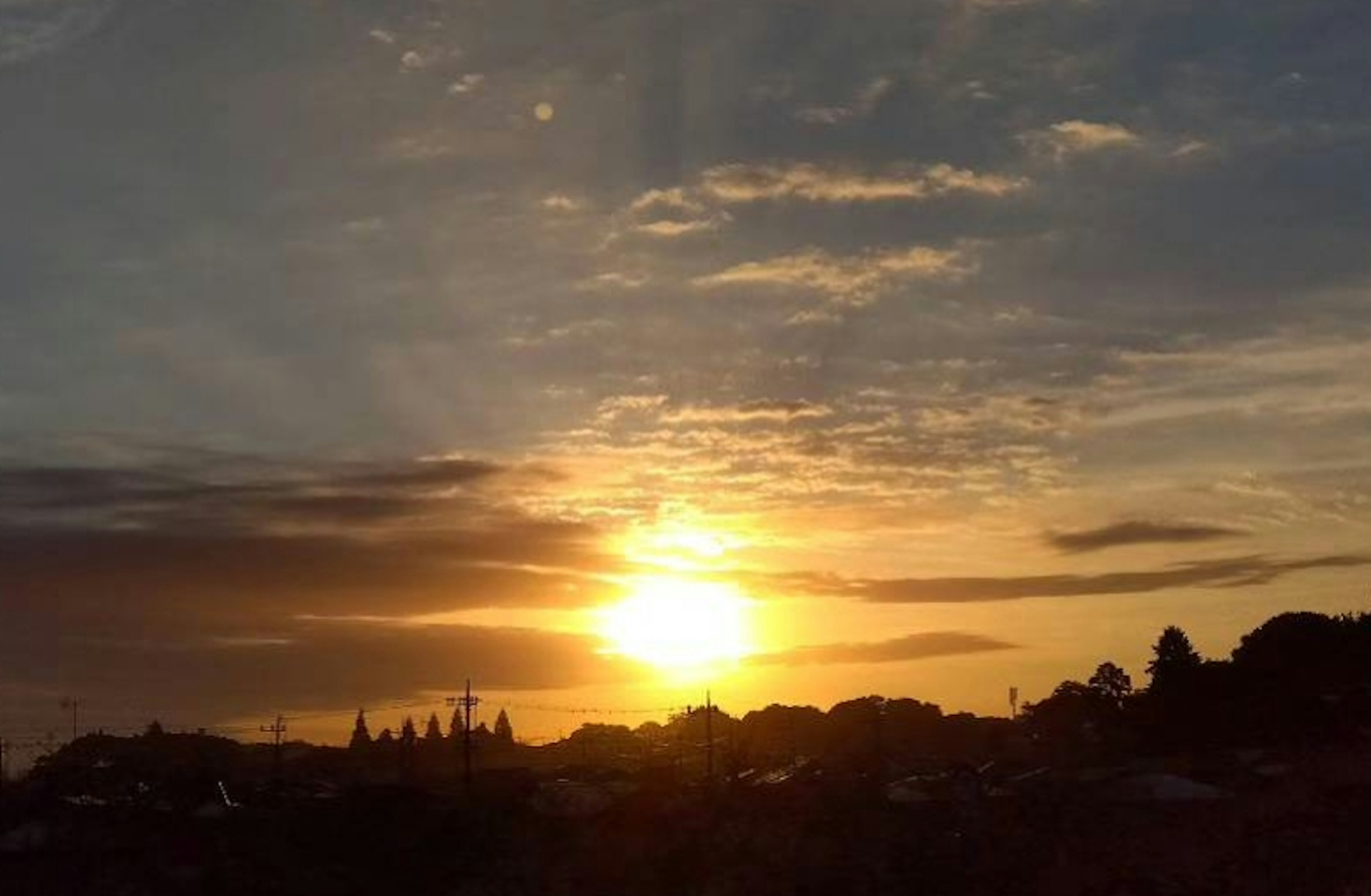 Sebuah matahari terbenam yang indah dengan nuansa oranye dan kuning lembut, membayangkan pohon-pohon dan bangunan