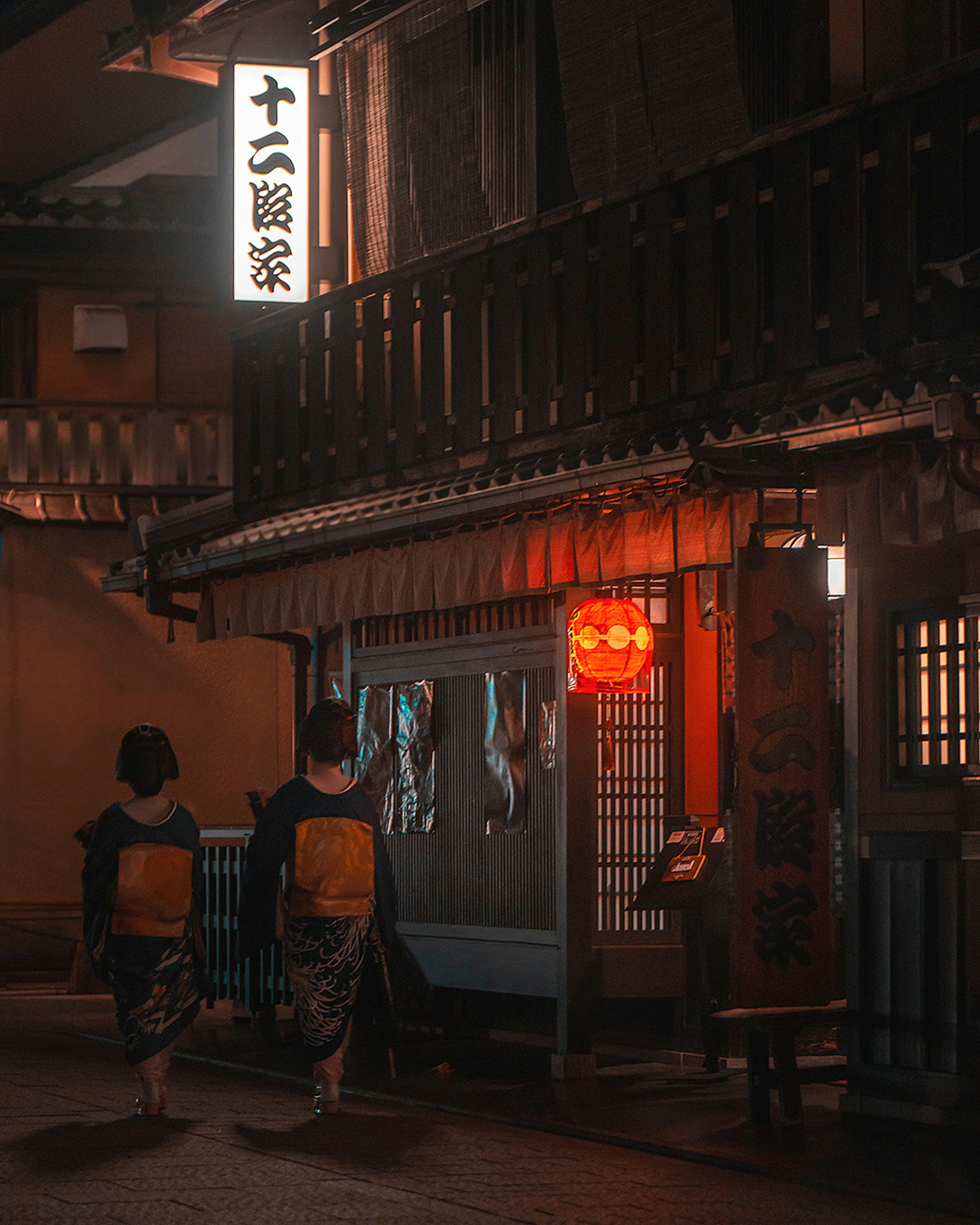 Two women walking in a traditional street at night wearing kimonos under a red lantern