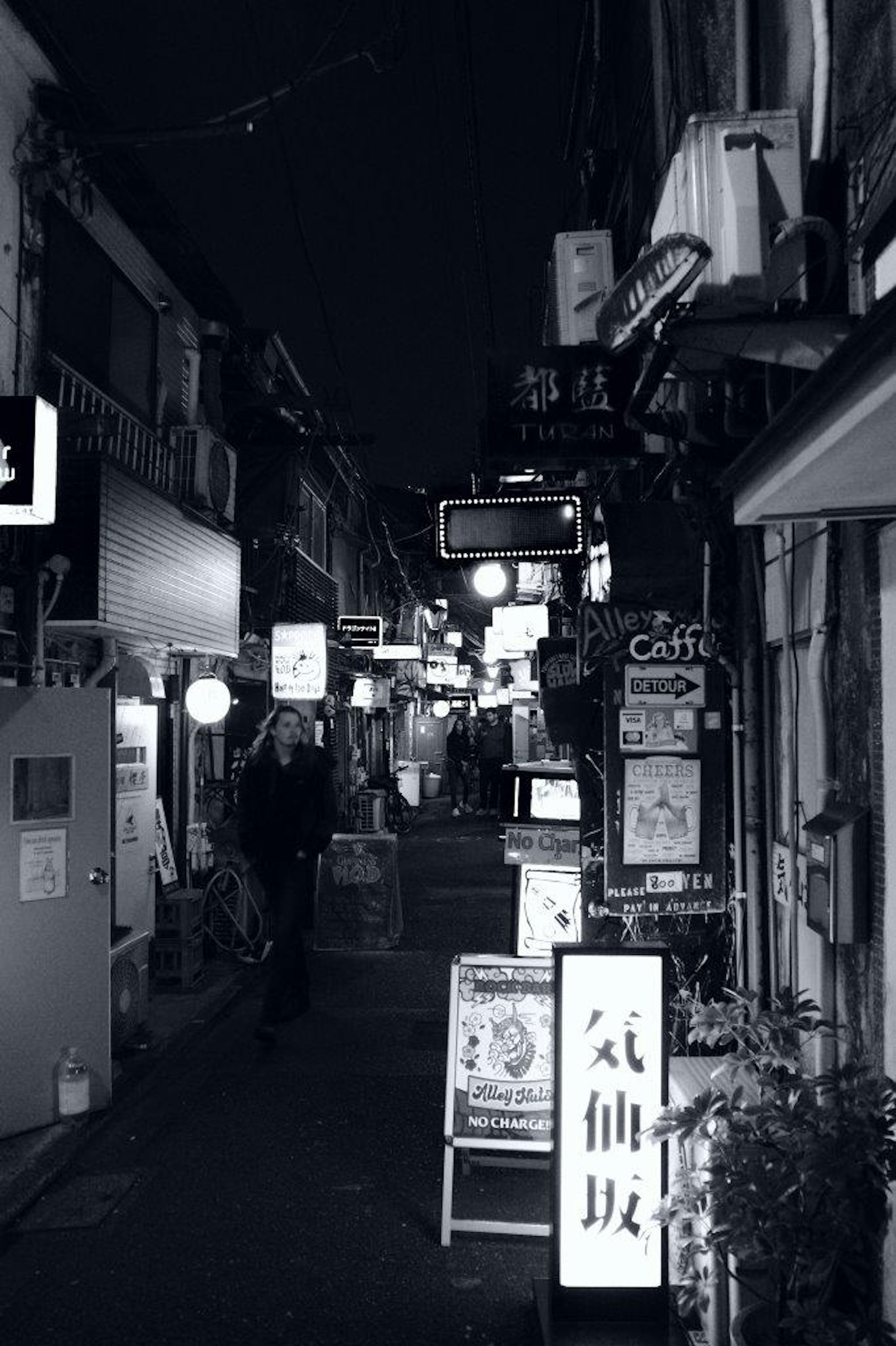 Callejón estrecho de noche con letreros de restaurantes iluminados y personas