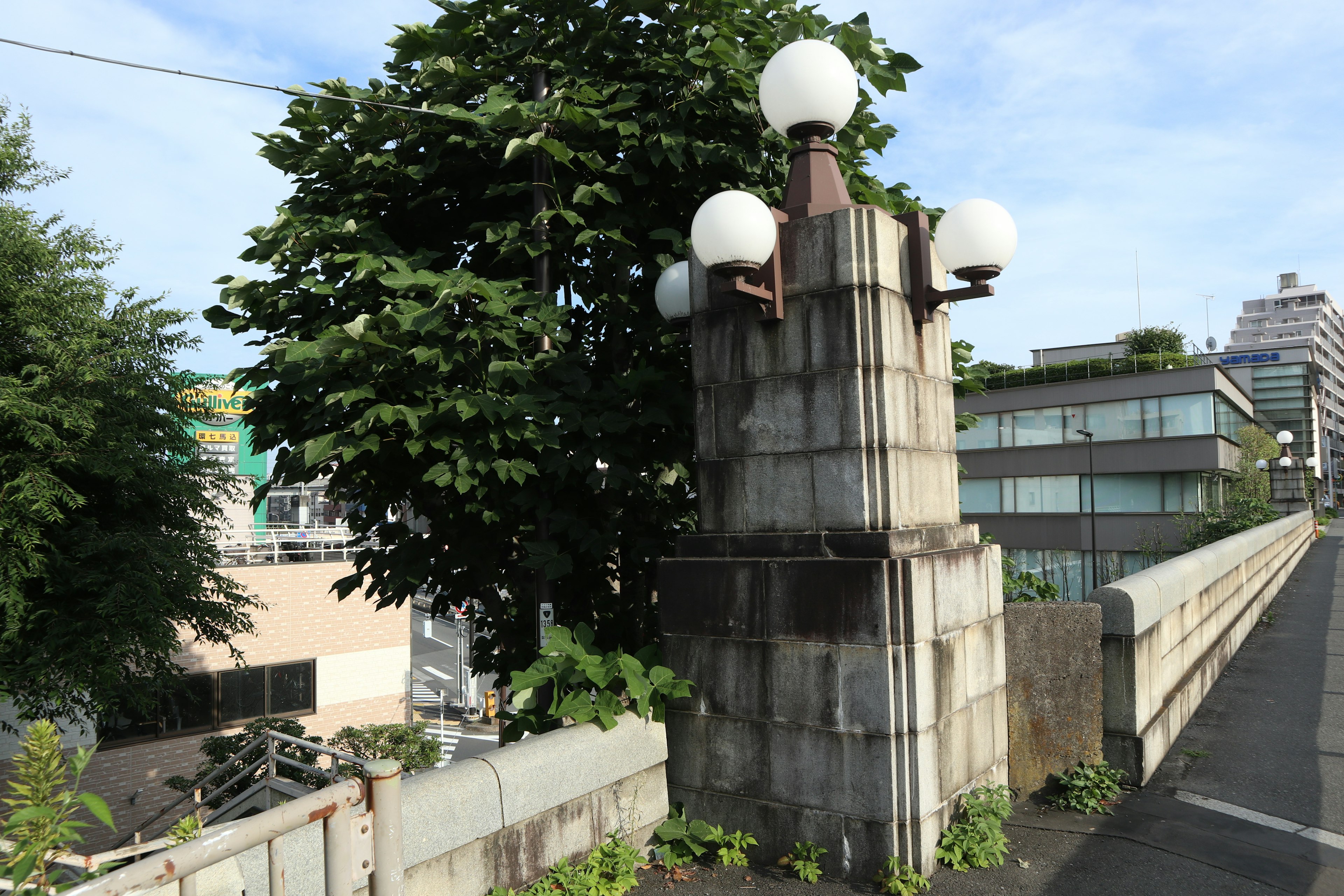 Steinerne Laternenstruktur auf einer Brücke mit Grün und modernen Gebäuden im Hintergrund