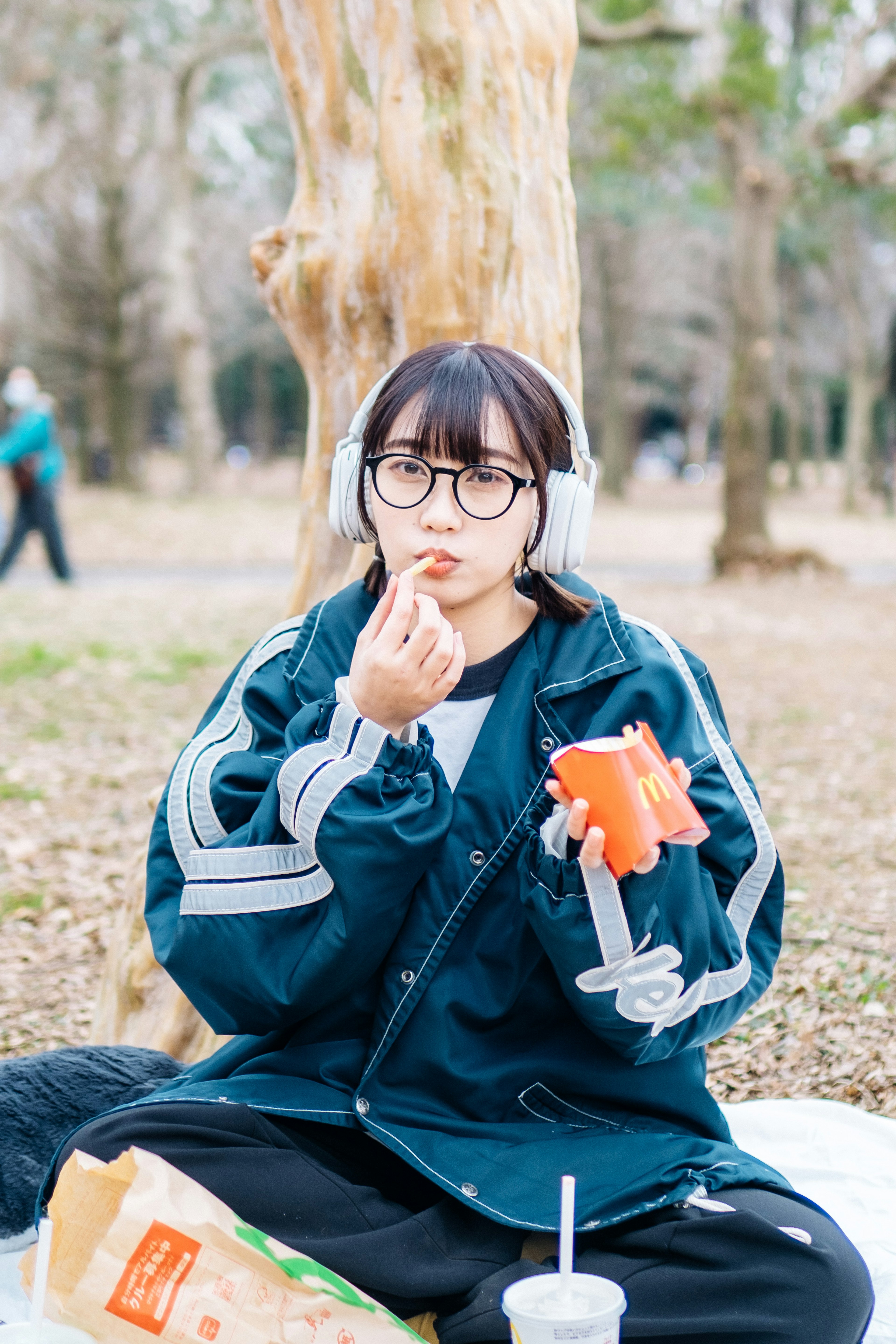 Donna che mangia snack in un parco indossando cuffie e occhiali vestita con pantaloni neri e giacca blu