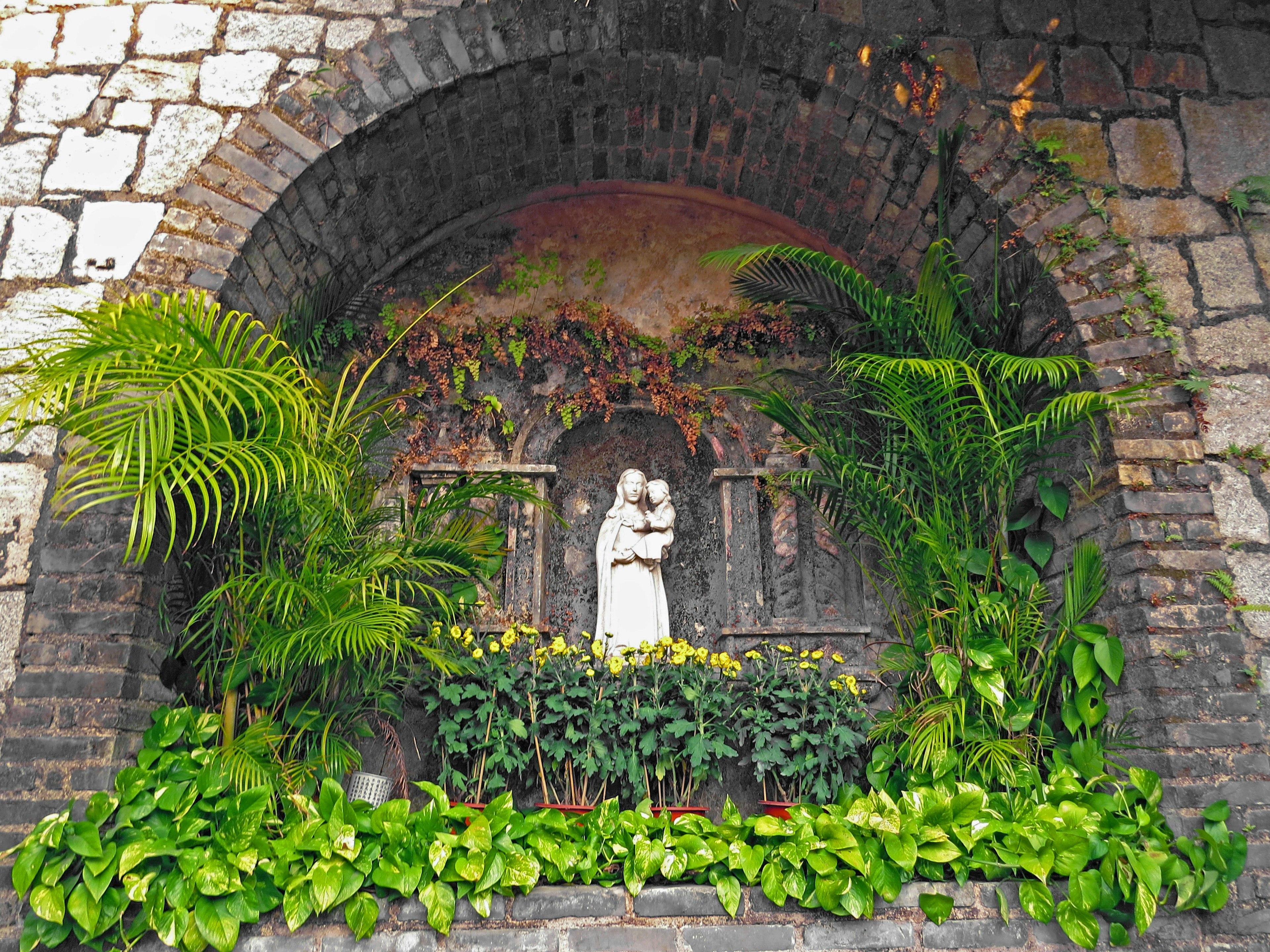 Nicho en arco con una estatua de la Virgen María rodeada de vegetación exuberante