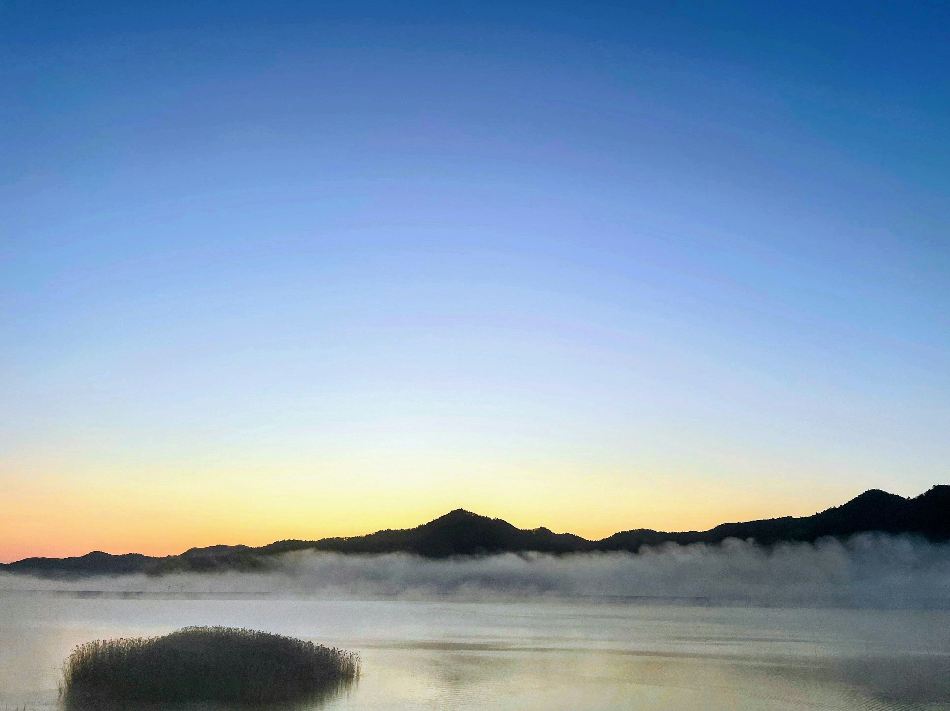 Ein ruhiger See umgeben von Nebel und Bergen unter einem lebhaften Himmel