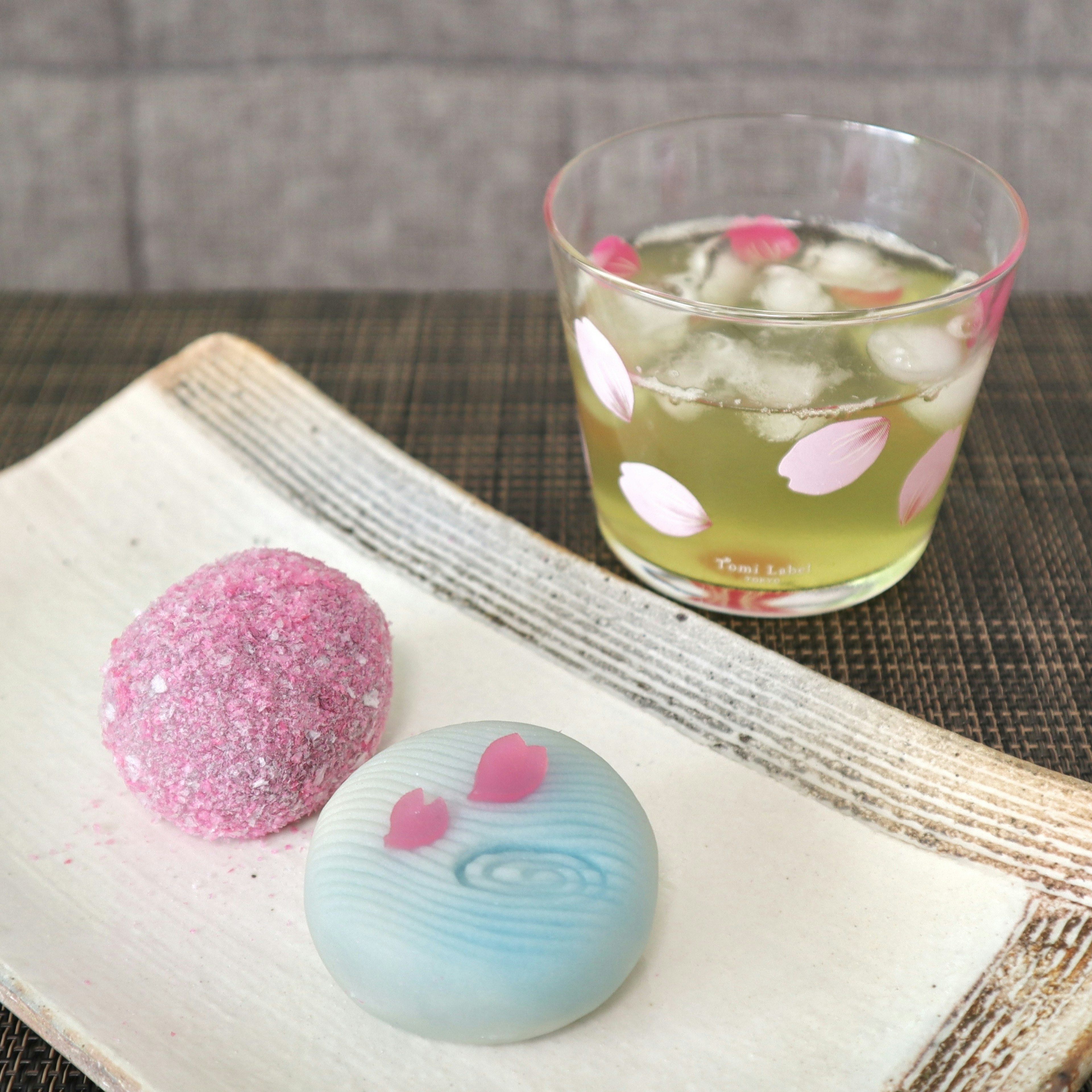 Dua wagashi berwarna pastel dan minuman dalam gelas bermotif sakura