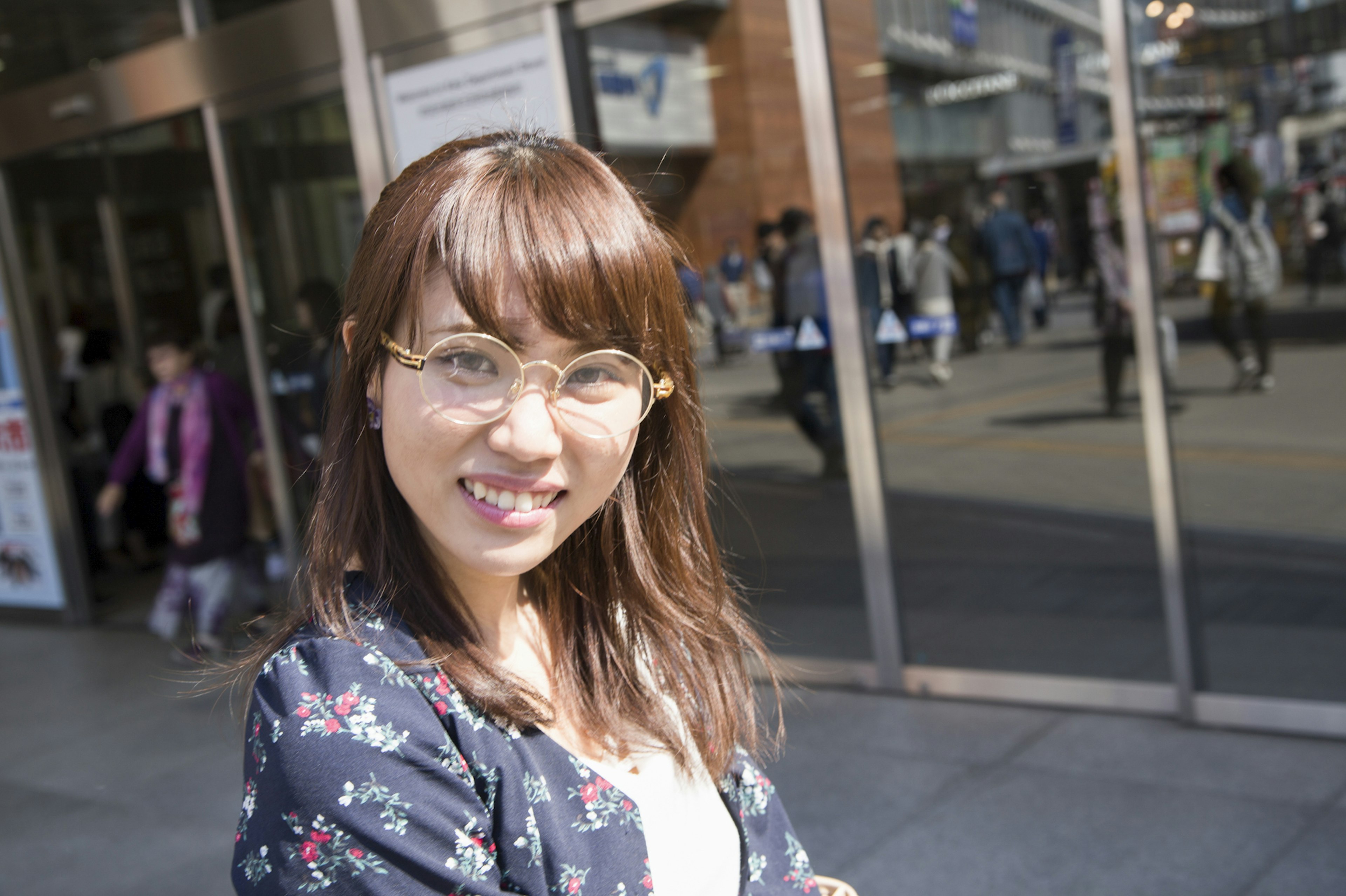 A woman wearing glasses smiles with a bustling street and people in the background