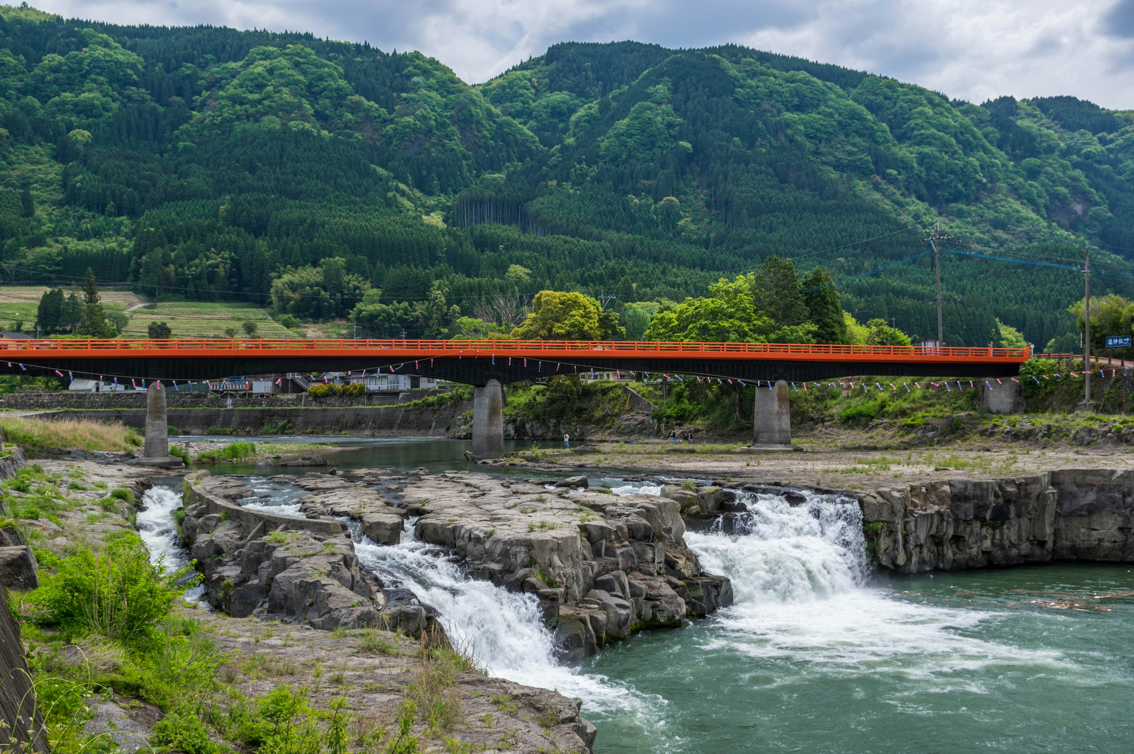 Malersicher Blick auf eine rote Brücke über einen Wasserfall mit üppigen grünen Bergen im Hintergrund