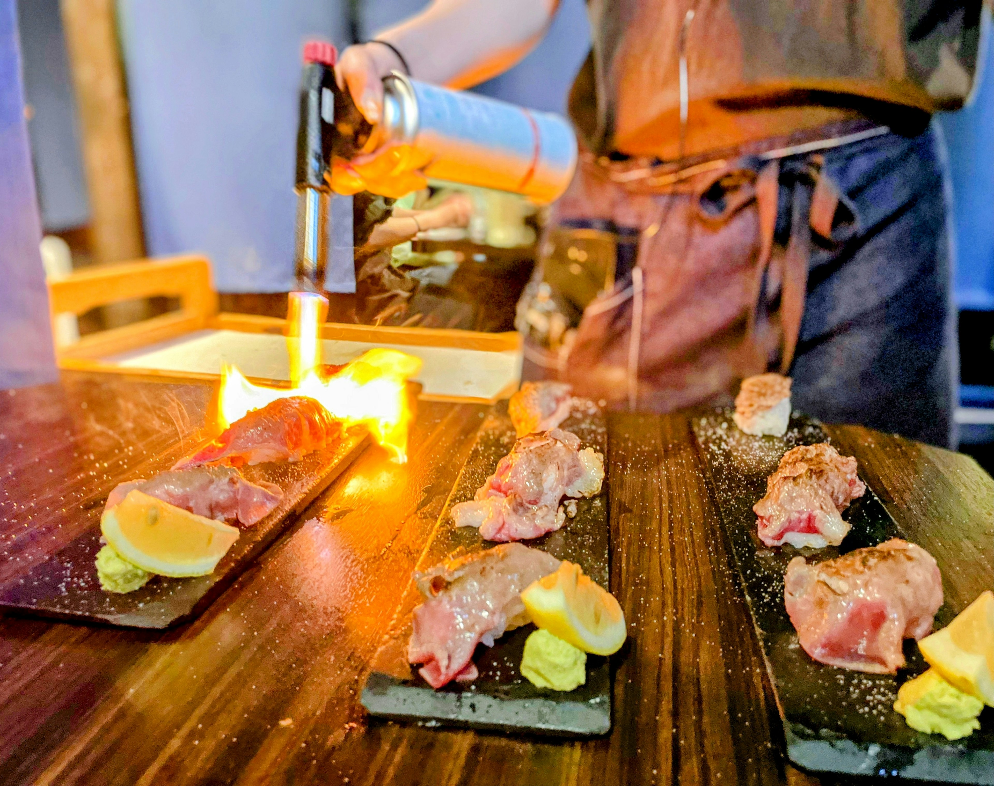 Chef searing meat with a torch on a wooden table with lemon and wasabi